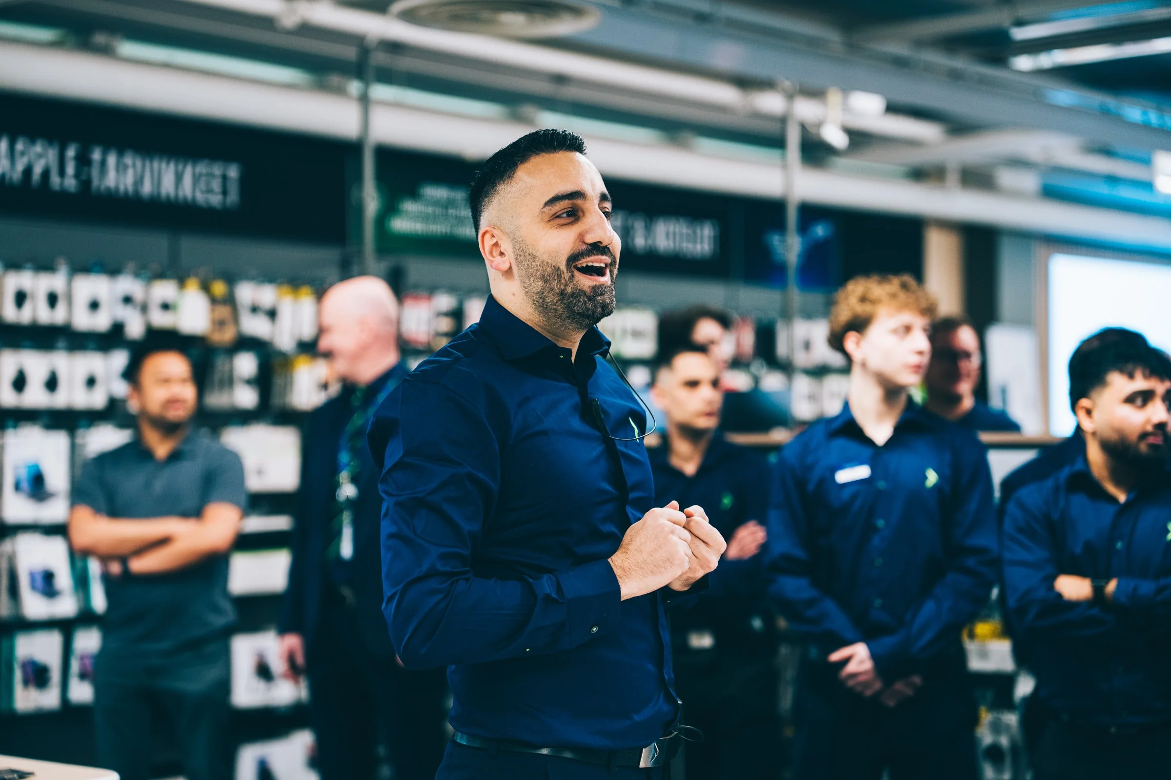 A man with dark hair and a beard wearing a dark blue shirt speaking or presenting to a group in a retail store with shelves of electronics and appliances in the background.