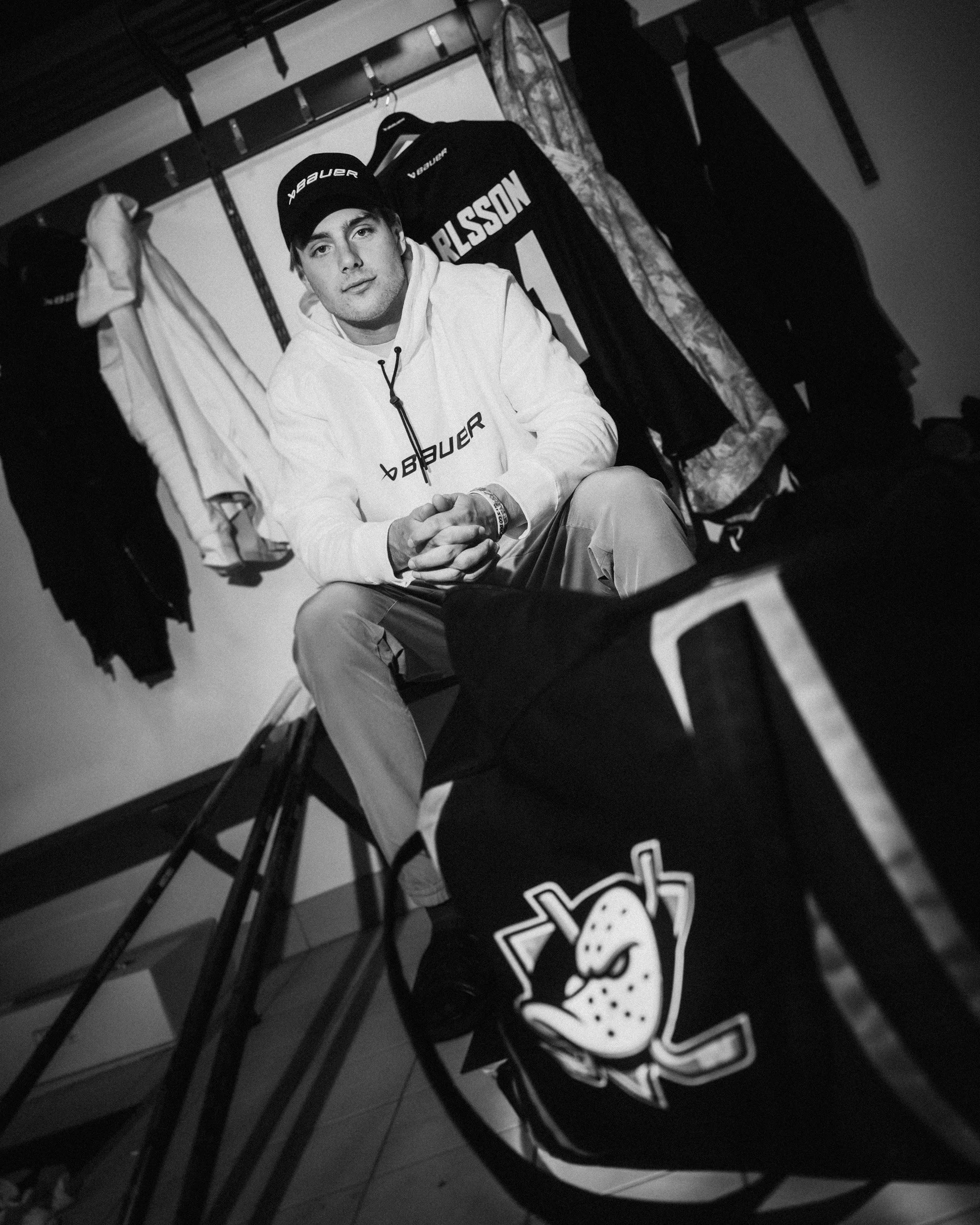 A young man sitting on a chair in a locker room. He is wearing a white hoodie with the word 'BAUER' on it and a black beanie hat. Behind him, there are hockey jerseys on hangers, including one with the name 'CARLSSON' and the number 51. The photo is 