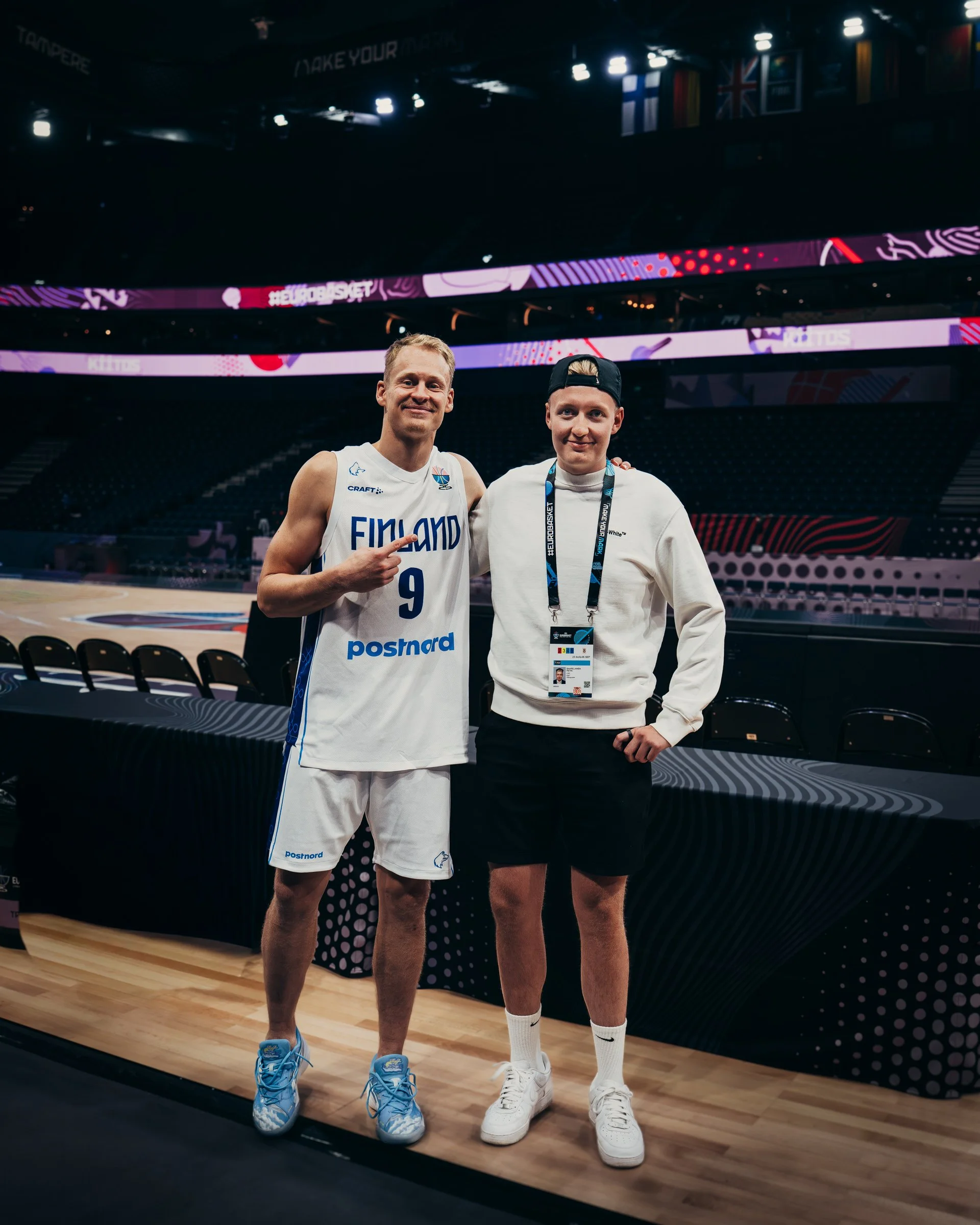 Two young men standing on a basketball court, one wearing a Finland basketball uniform with number 9, pointing and smiling, the other in a white sweatshirt, black shorts, and a backwards cap, posing with an accreditation badge, in an empty arena.
