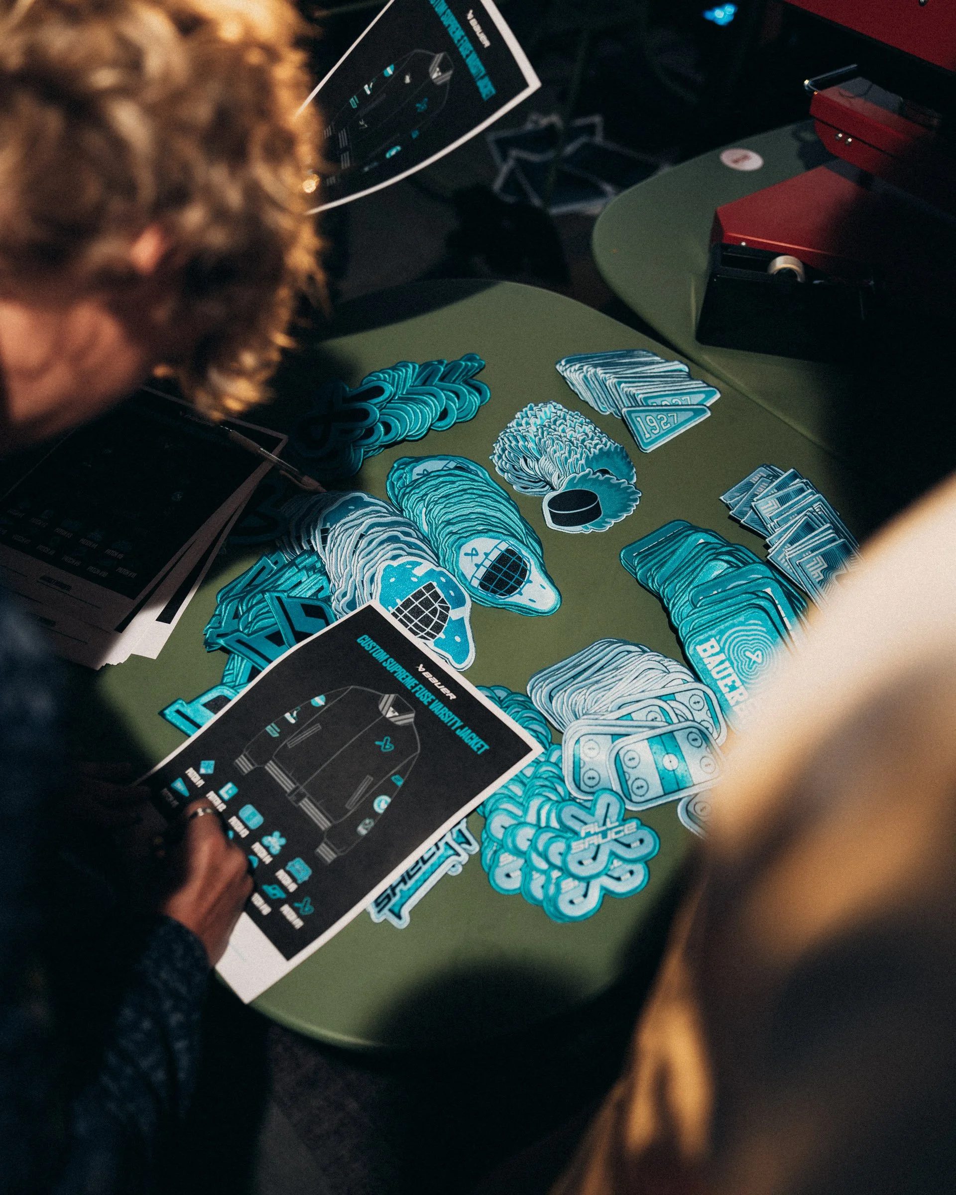 Close-up of a green table with blue and white stickers and patches featuring designs and text such as '1987' and 'CUSTOM SUPREME + FUSE VARSITY JACKETS.' A person's hand is holding a flooded card. In the background, there are instructions and a devic