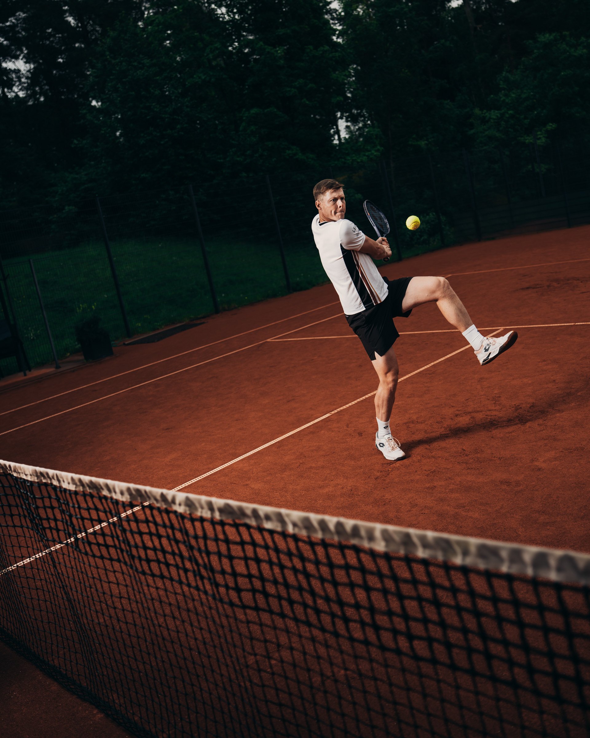 A male tennis player in a white shirt and black shorts hits a tennis ball on an outdoor court surrounded by green trees.