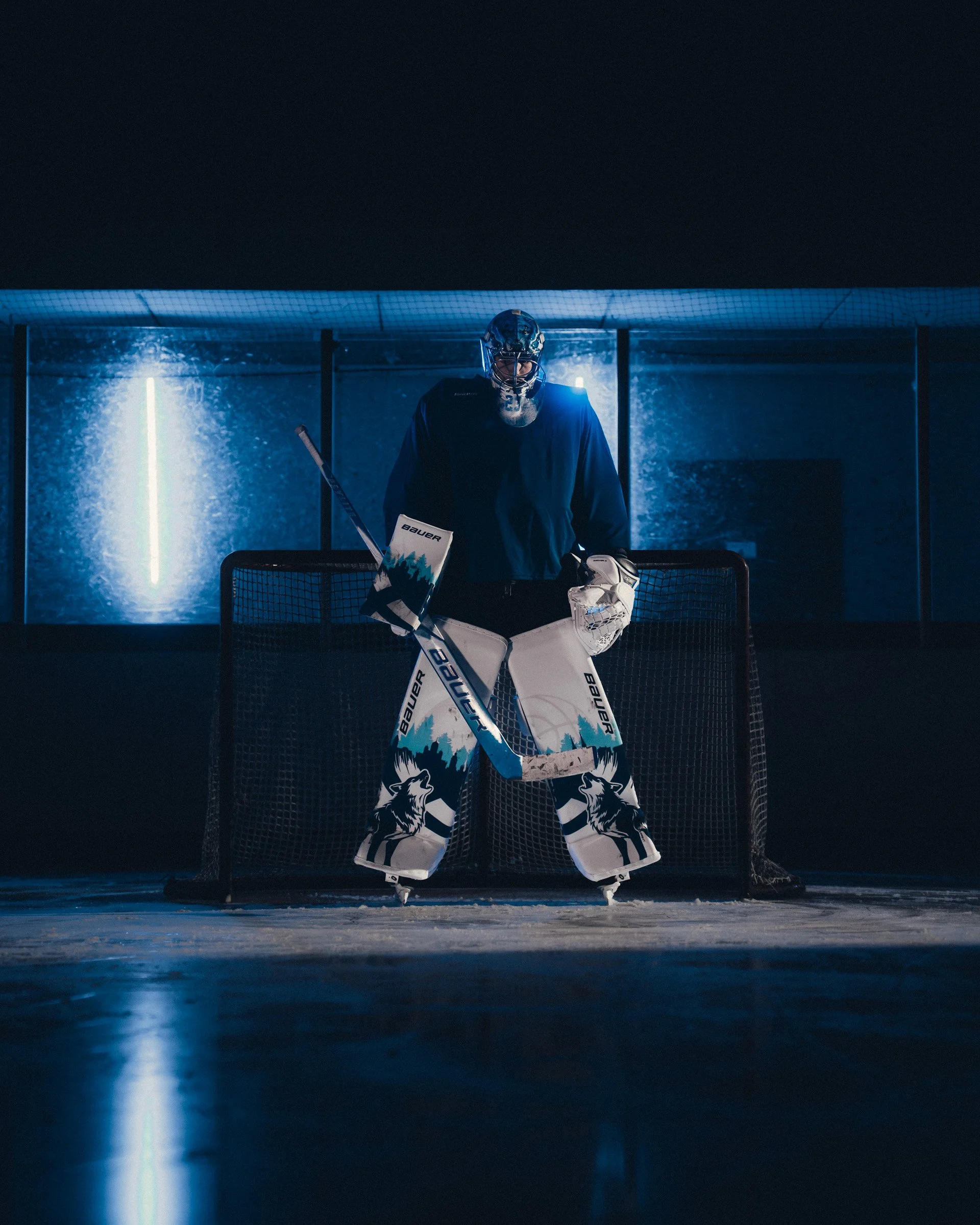 Ice hockey goalie standing in front of the net on an ice rink, illuminated with blue lighting.