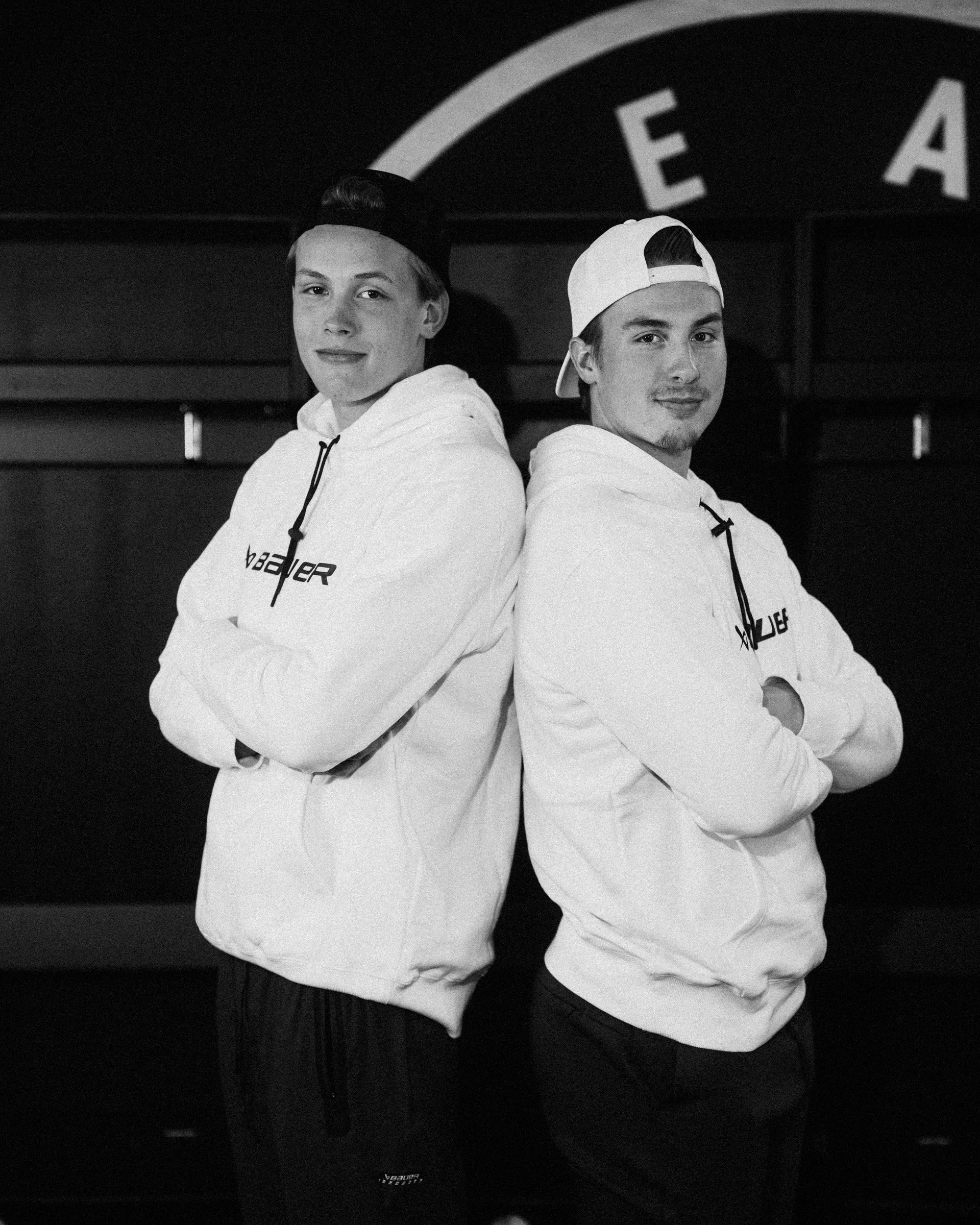 Two young men standing back to back with arms crossed, wearing white hoodies and baseball caps backward, with a dark background featuring a circular logo with the letters 'E A'.