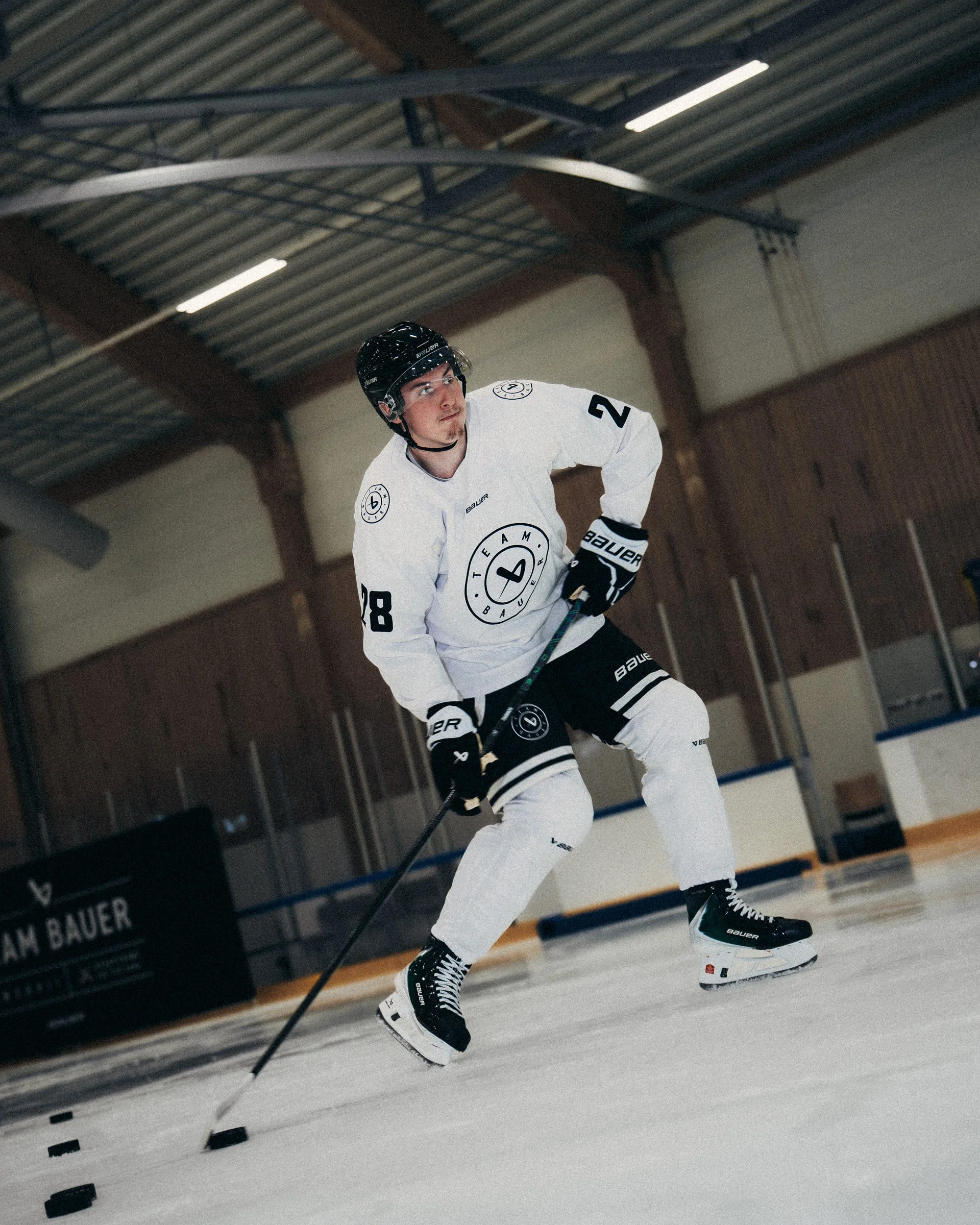 An ice hockey player in a white jersey with black gloves and black helmet skating on indoor ice rink. The jersey has a circular logo, the number 28, and the brand Bauer. The player is holding a hockey stick and looking ahead with focus.