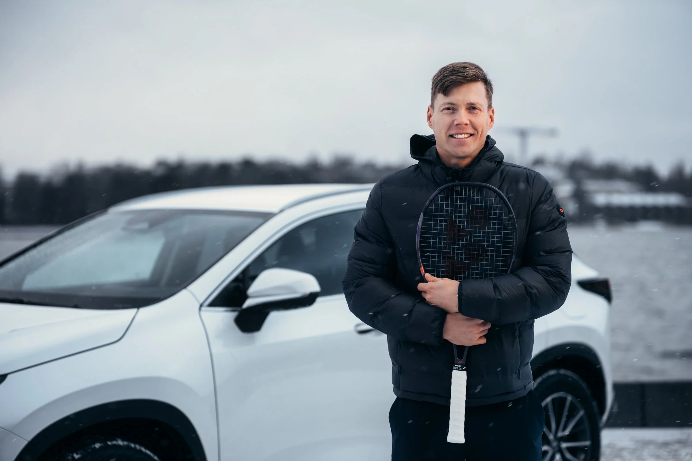A young man in a black jacket holding a tennis racket stands outside near a white SUV on a cloudy day, with a body of water and land in the background.