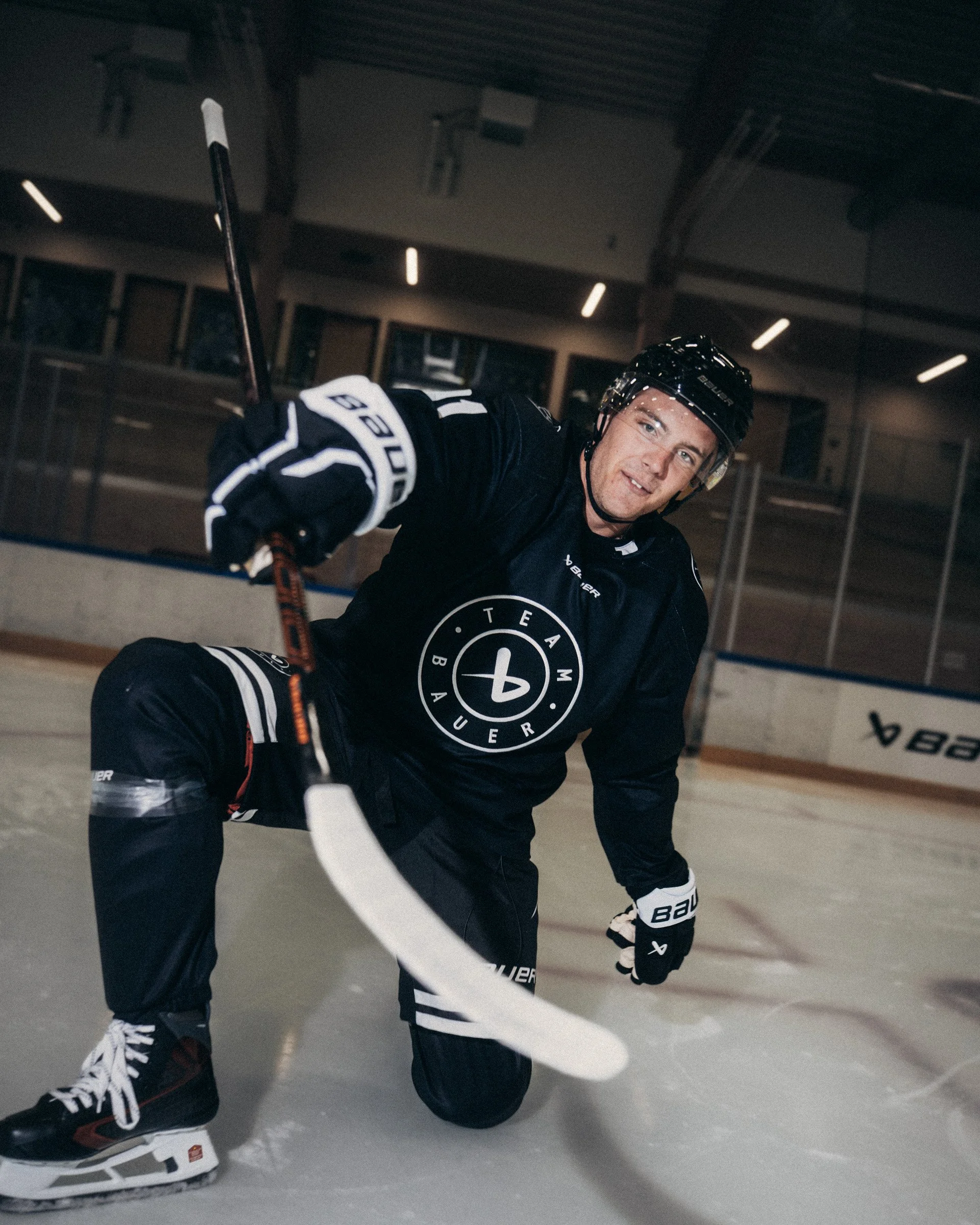 A male ice hockey player in black jersey and helmet kneeling on ice rink, smiling, with hockey stick in hand, wearing Bauer gloves and skate boots.