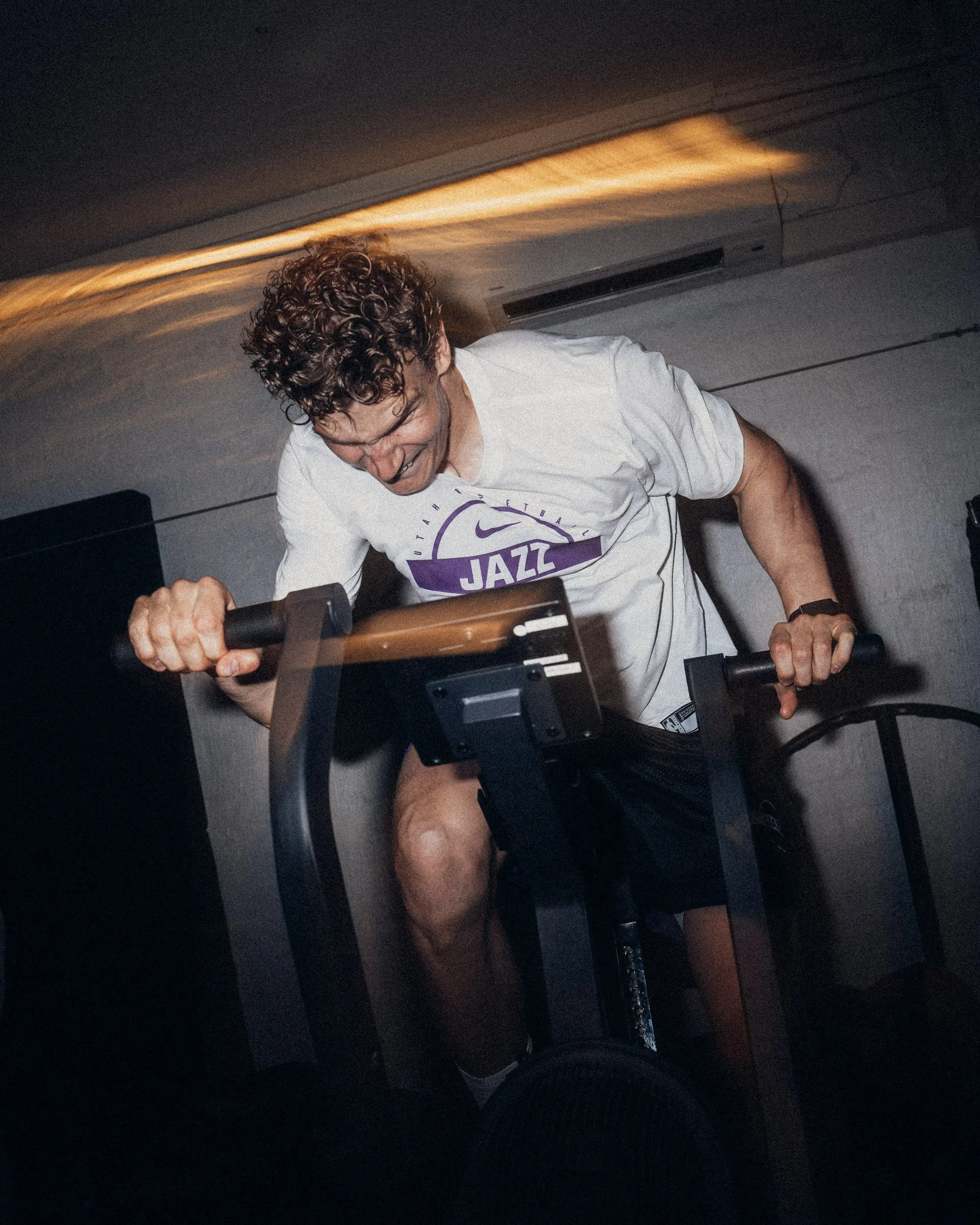 A man with curly hair is riding a stationary exercise bike in a dimly lit room. He is wearing a white T-shirt with purple text and is smiling or laughing.