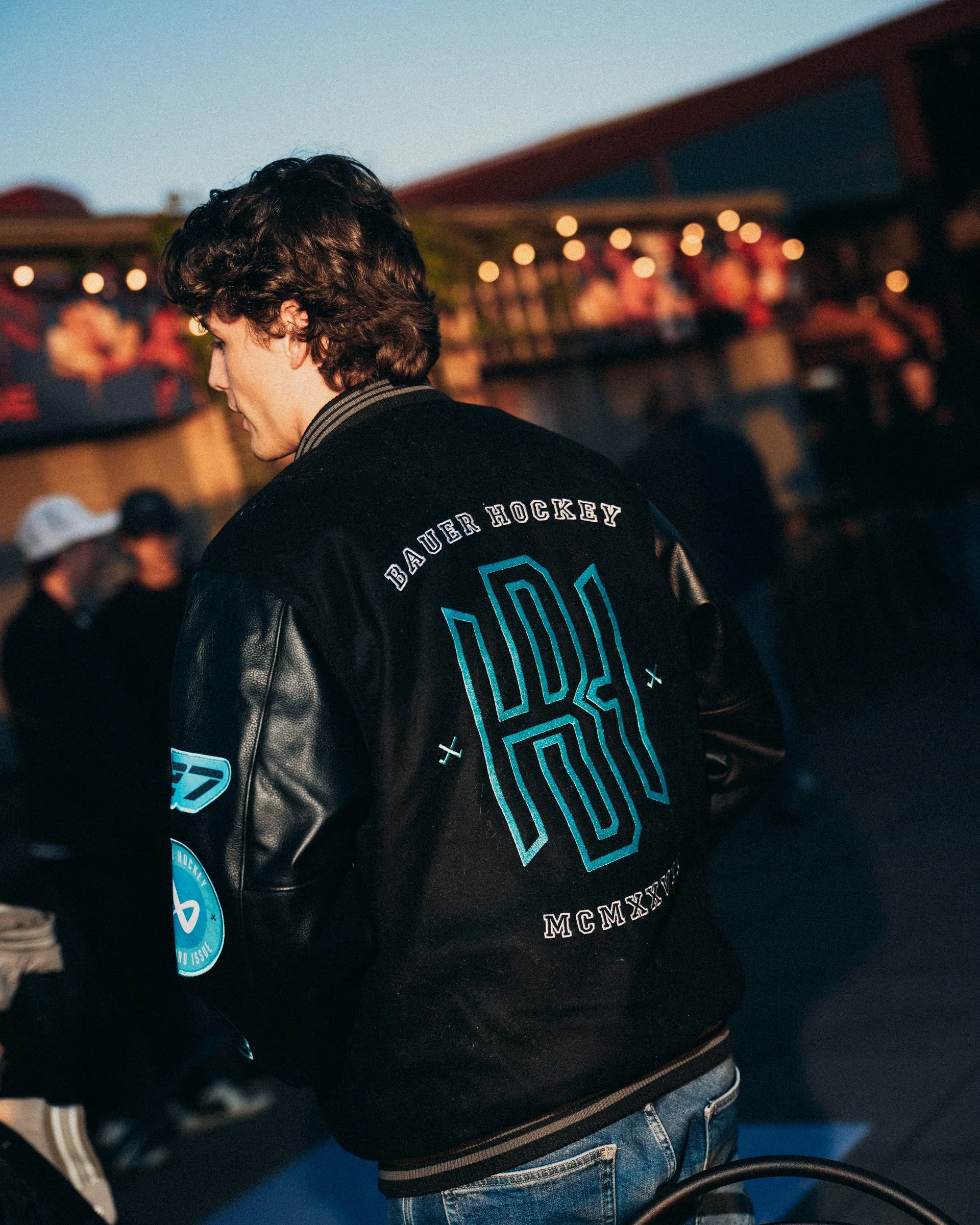 Back of a young man with wavy dark hair, wearing a black hockey jacket with blue patches, standing in a crowd at dusk.