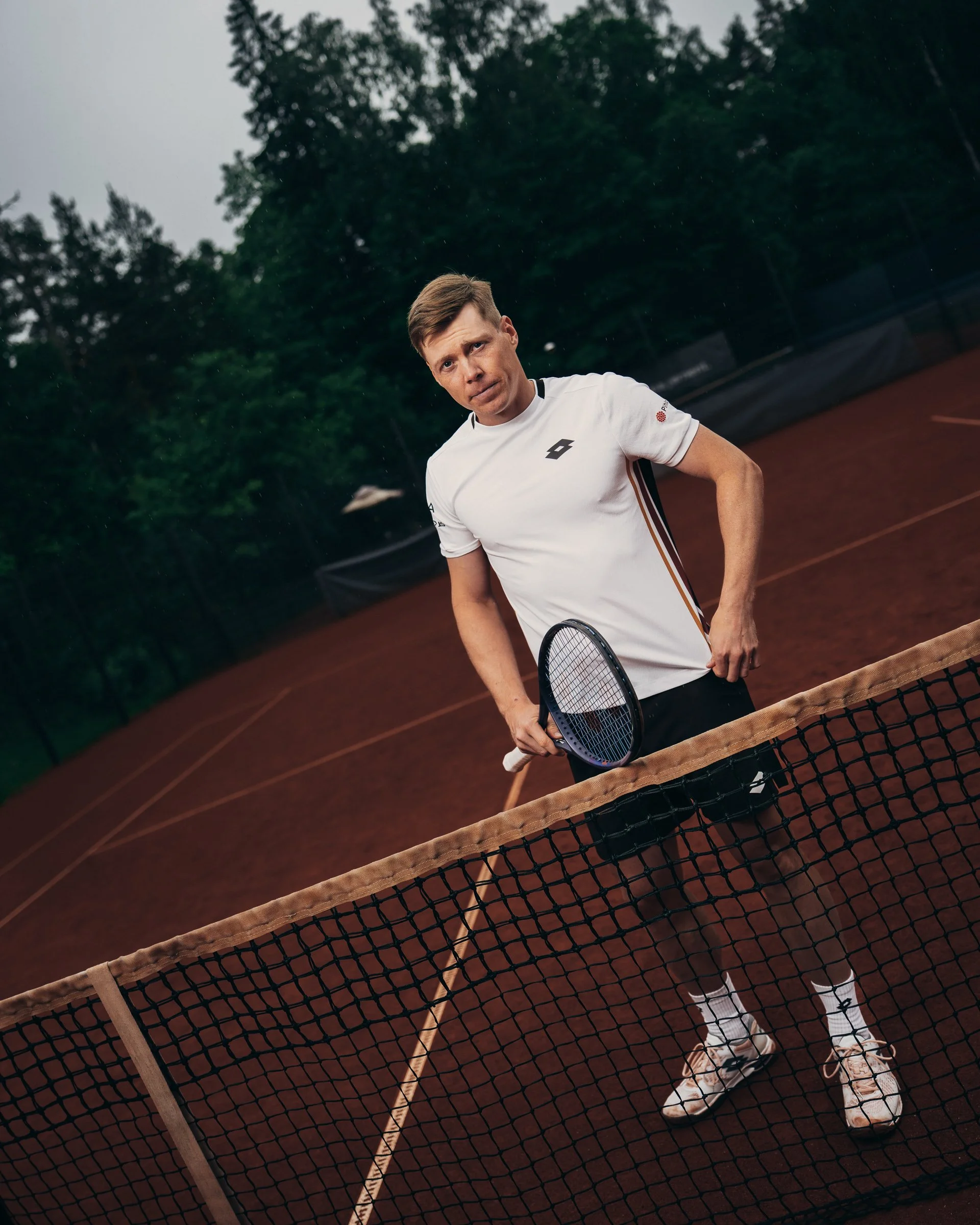 A man holding a tennis racket on a tennis court, with a net in the foreground and green trees in the background, overcast sky.