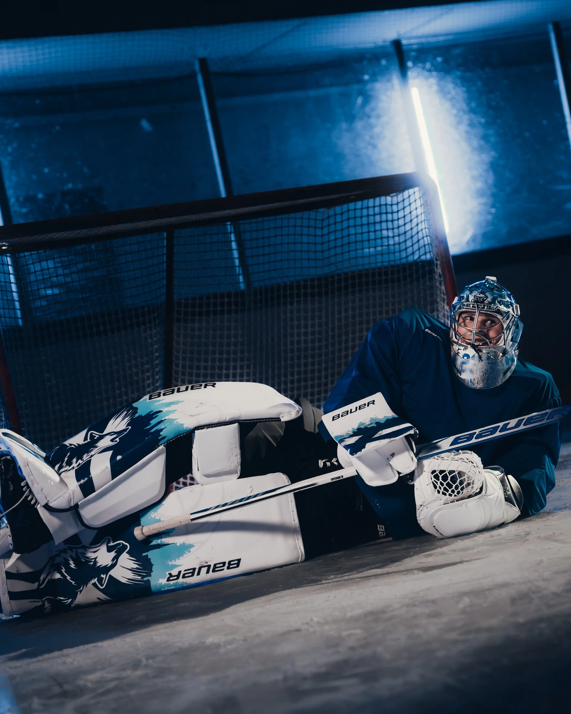 An ice hockey goalkeeper in a blue uniform and protective mask is on the ice in front of the goal, wearing white goalie pads with wolf graphics and the brand name 'Bauer' visible, with a netted goalpost in the background.