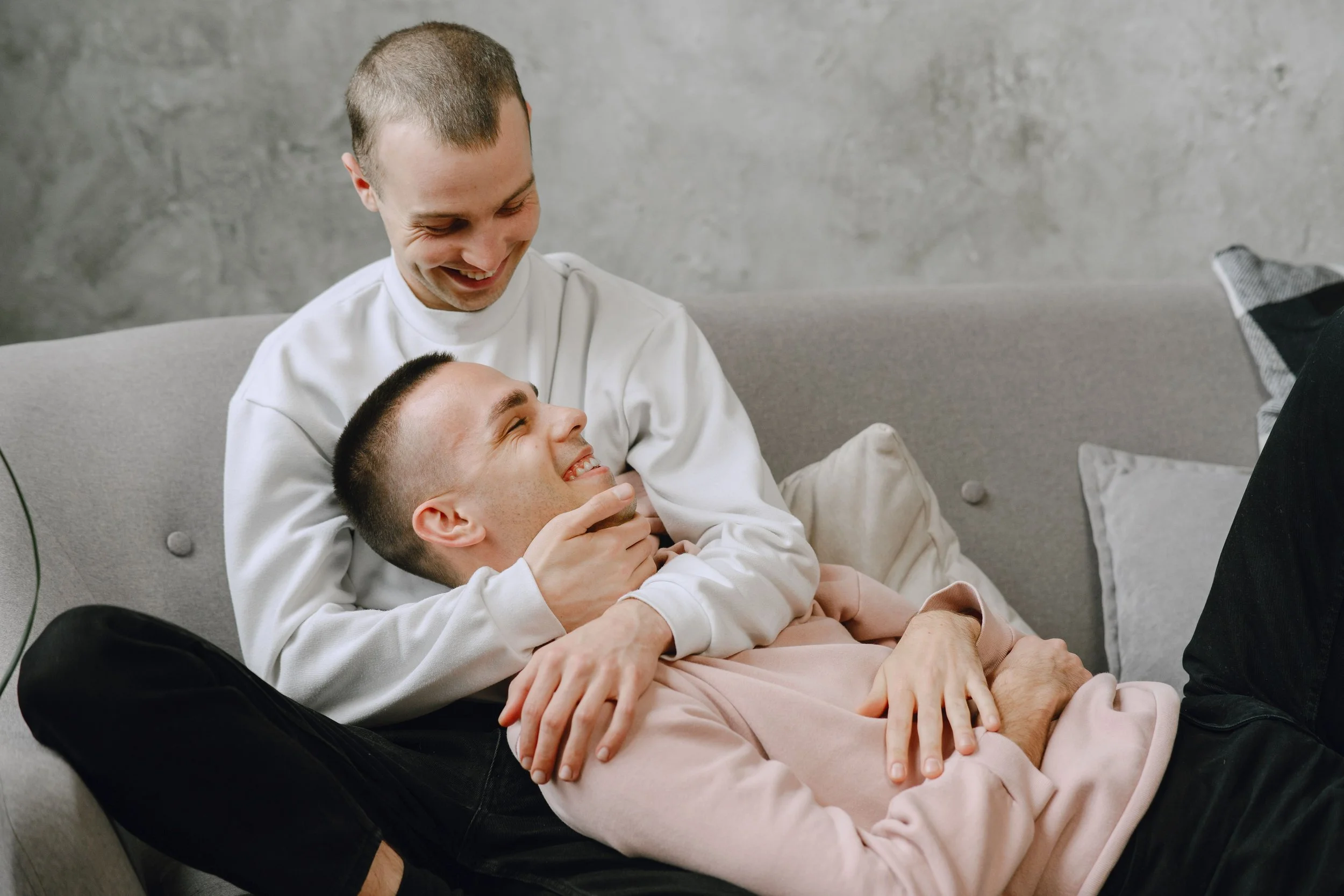 Two young men relaxing together on a couch in a cozy living room, smiling and enjoying each other’s company.
