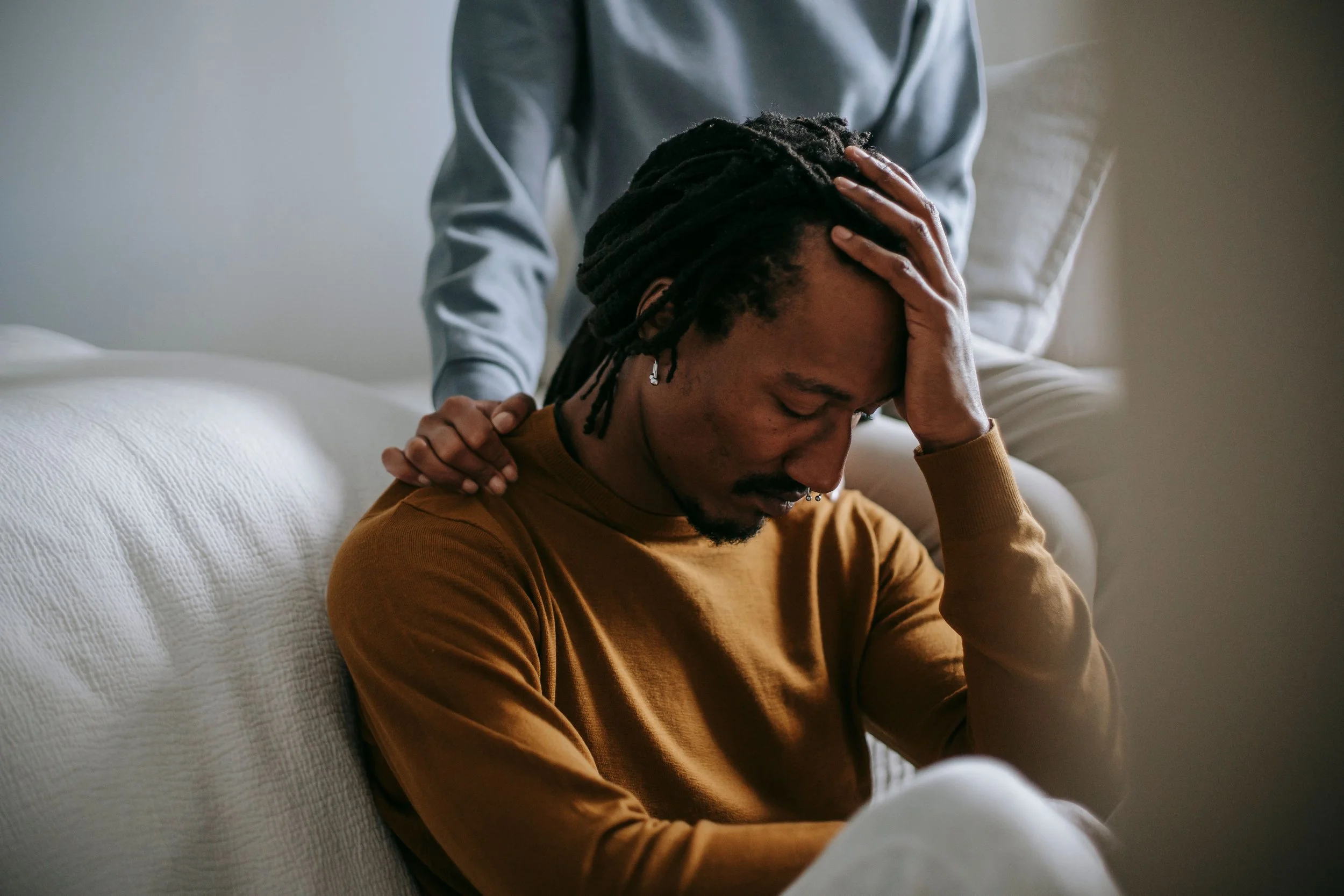 Man with dreadlocks sitting on a white couch, holding his head with one hand as another person gently rests a hand on his shoulder.