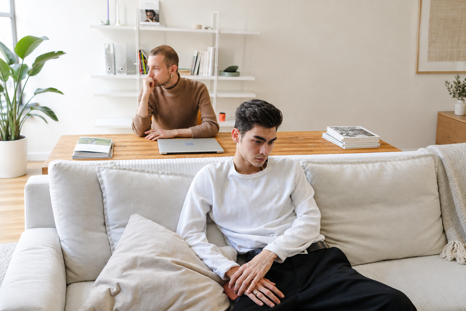 Two men in a living room, one sitting on a white couch while the other sits at a dining table with a laptop and magazines.