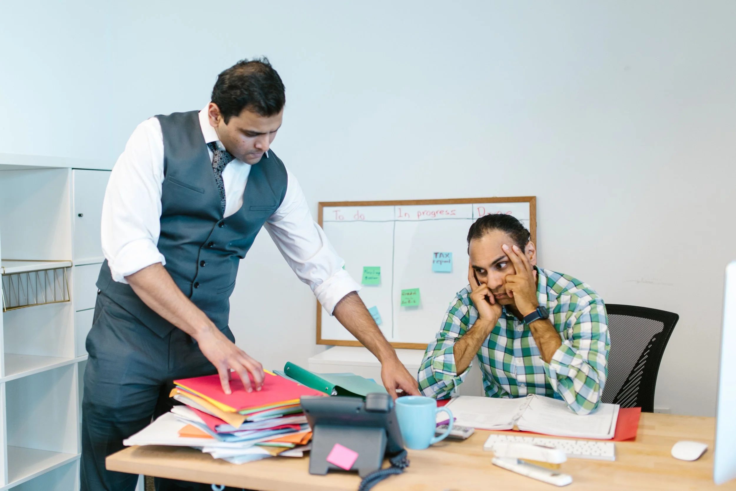 Two men are in an office, one standing and the other sitting. The man standing is dressed in a grey vest and white shirt, while the man sitting is wearing a checkered shirt, holding his head, looking stressed. The desk has a large pile of colorful folders, a phone, a blue mug, and scattered papers.