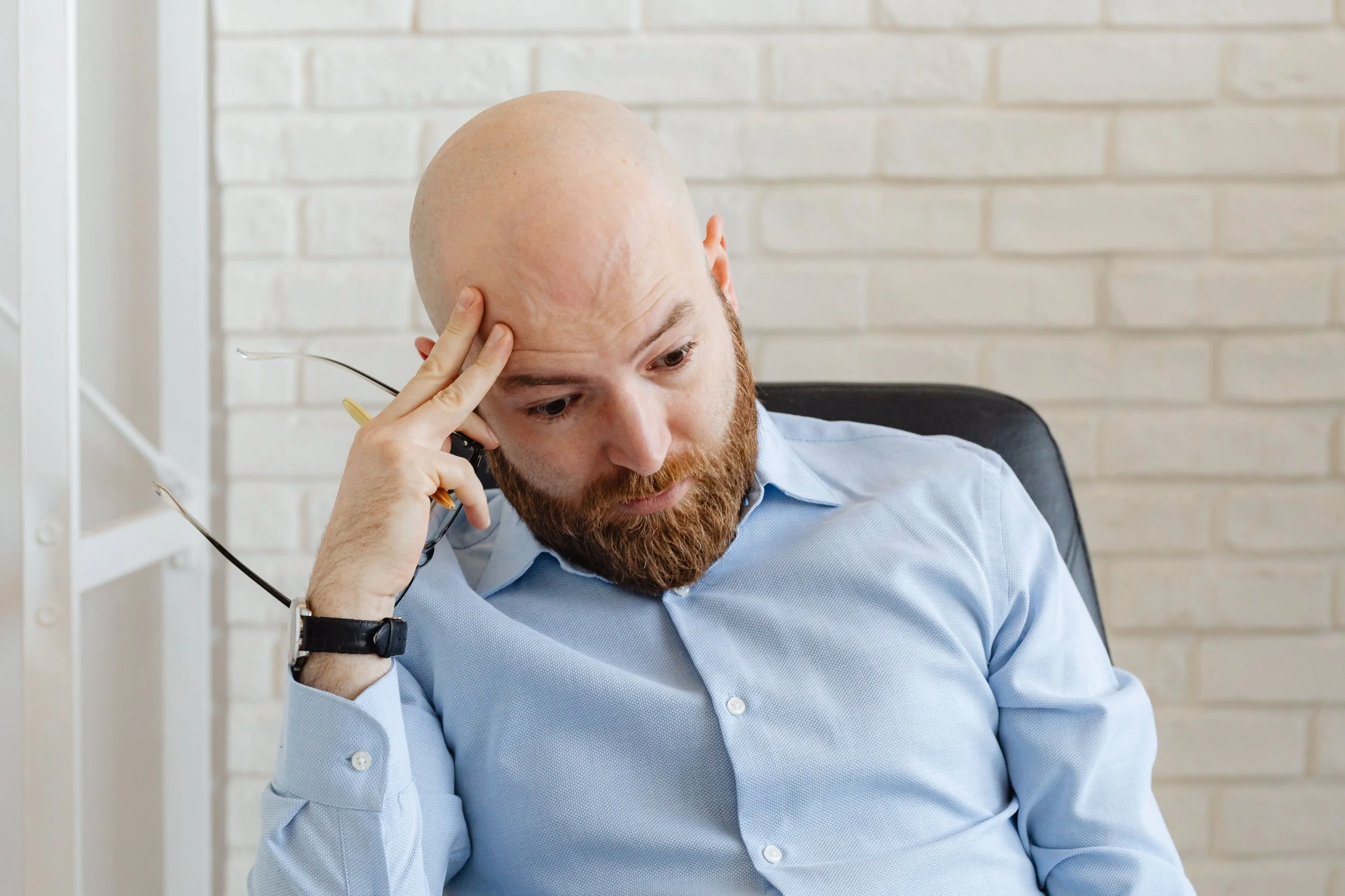 Bearded man in a light blue shirt sitting against a white brick wall, resting his head on his hand while holding glasses.
