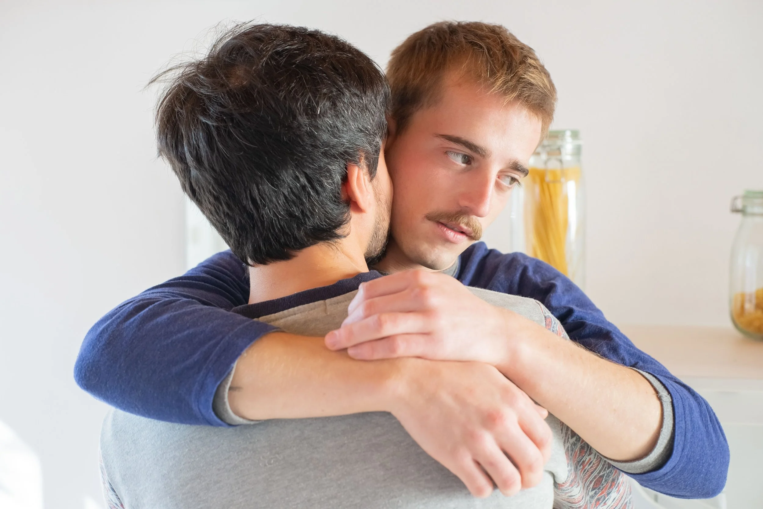 Young man with reddish hair hugging an older man, both smiling softly in a well-lit room.
