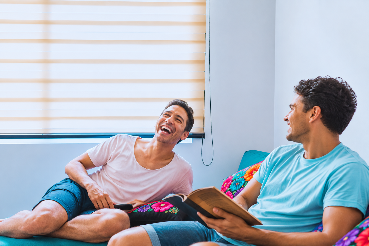 Two men sitting on a colorful floral couch, laughing and talking, with one holding a book and the other holding a TV remote in a bright room.