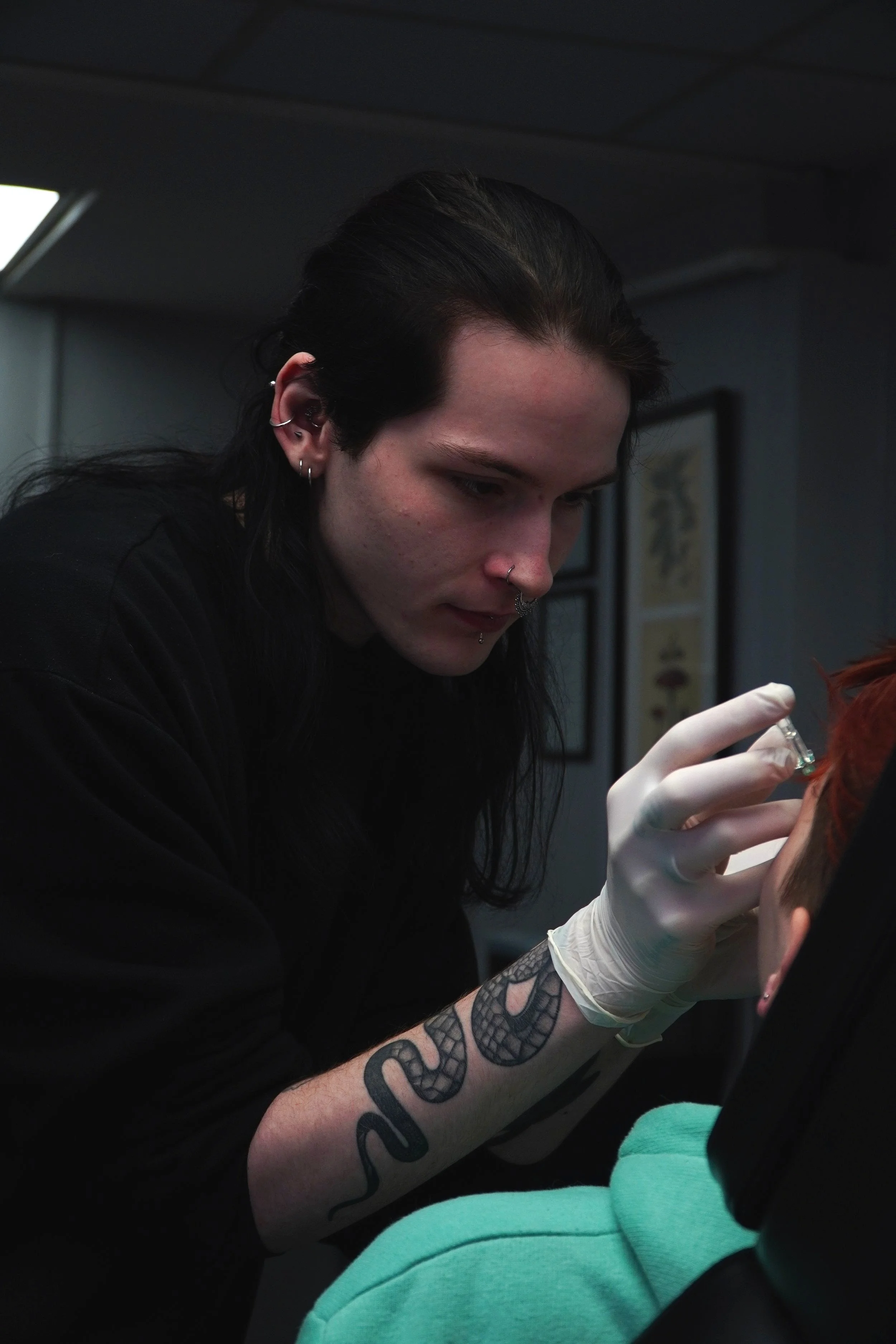 A tattooed woman with dark hair and multiple piercings, wearing gloves, is administering a tattoo to a person with red hair, who is wearing a green hoodie.