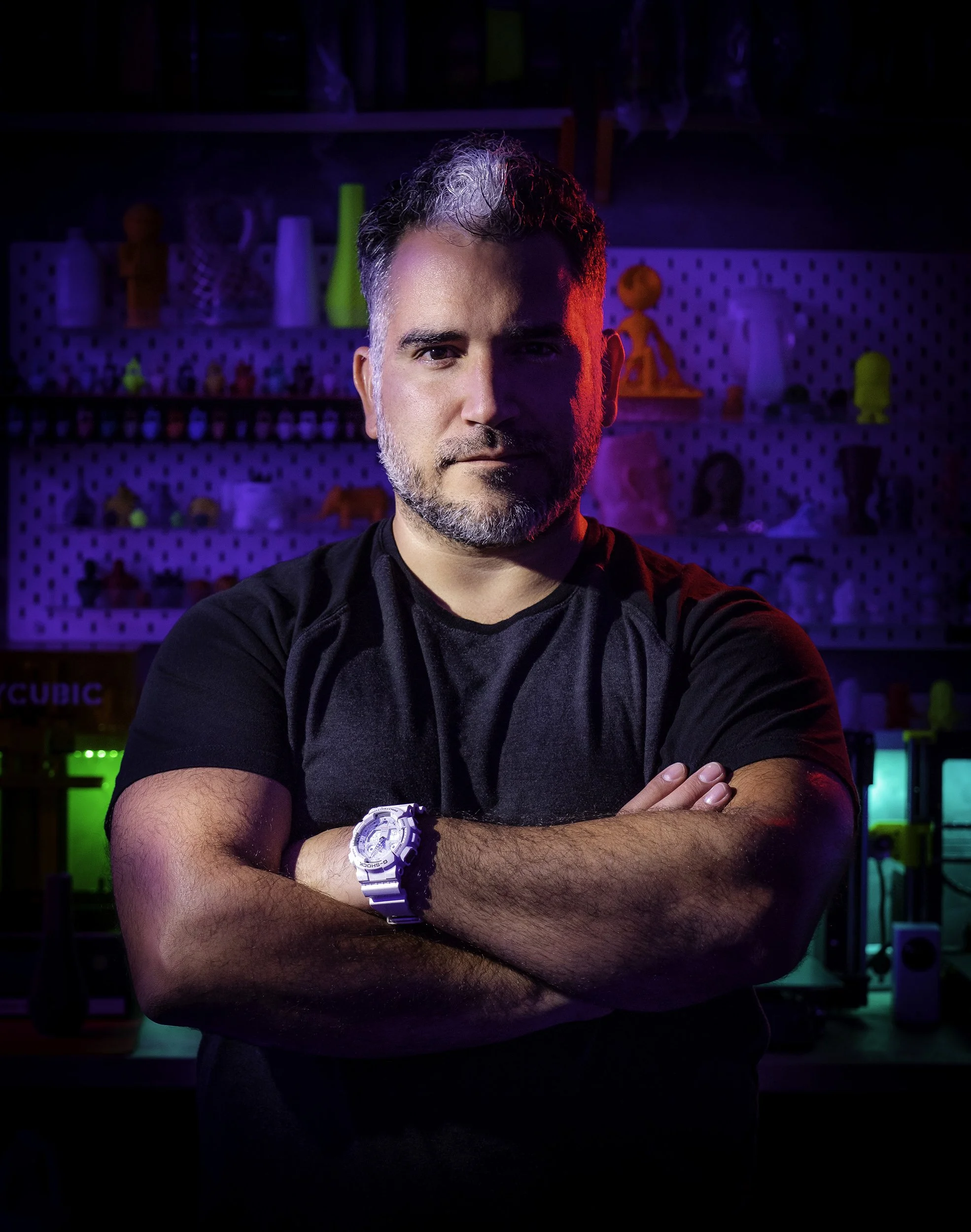 A middle-aged man with gray hair, beard, and mustache standing with arms crossed in a room with colorful shelves and toys in the background, illuminated with neon purple, pink, and blue lighting.