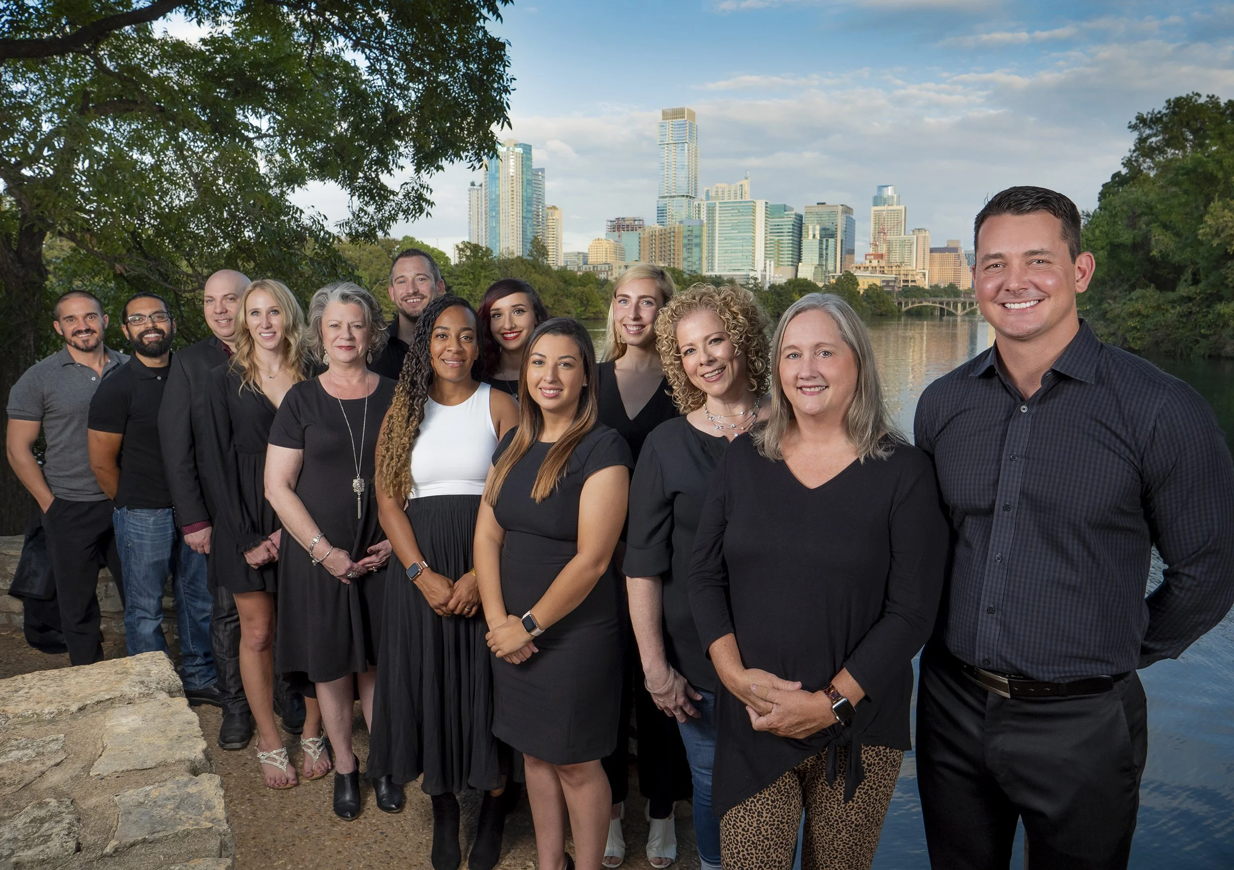A group of diverse professionals standing outdoors near a river with a city skyline in the background, during daytime.