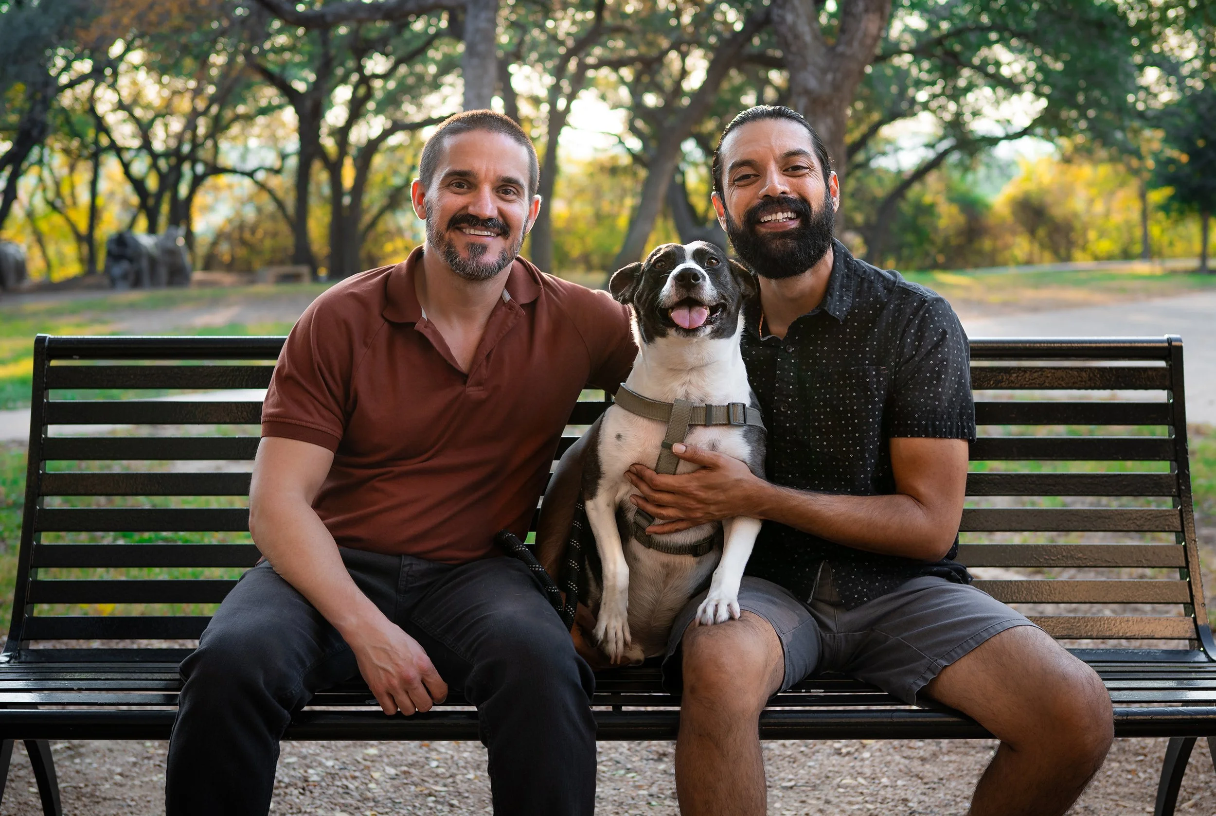 Two men sitting on a park bench with a dog between them, surrounded by trees with autumn foliage.