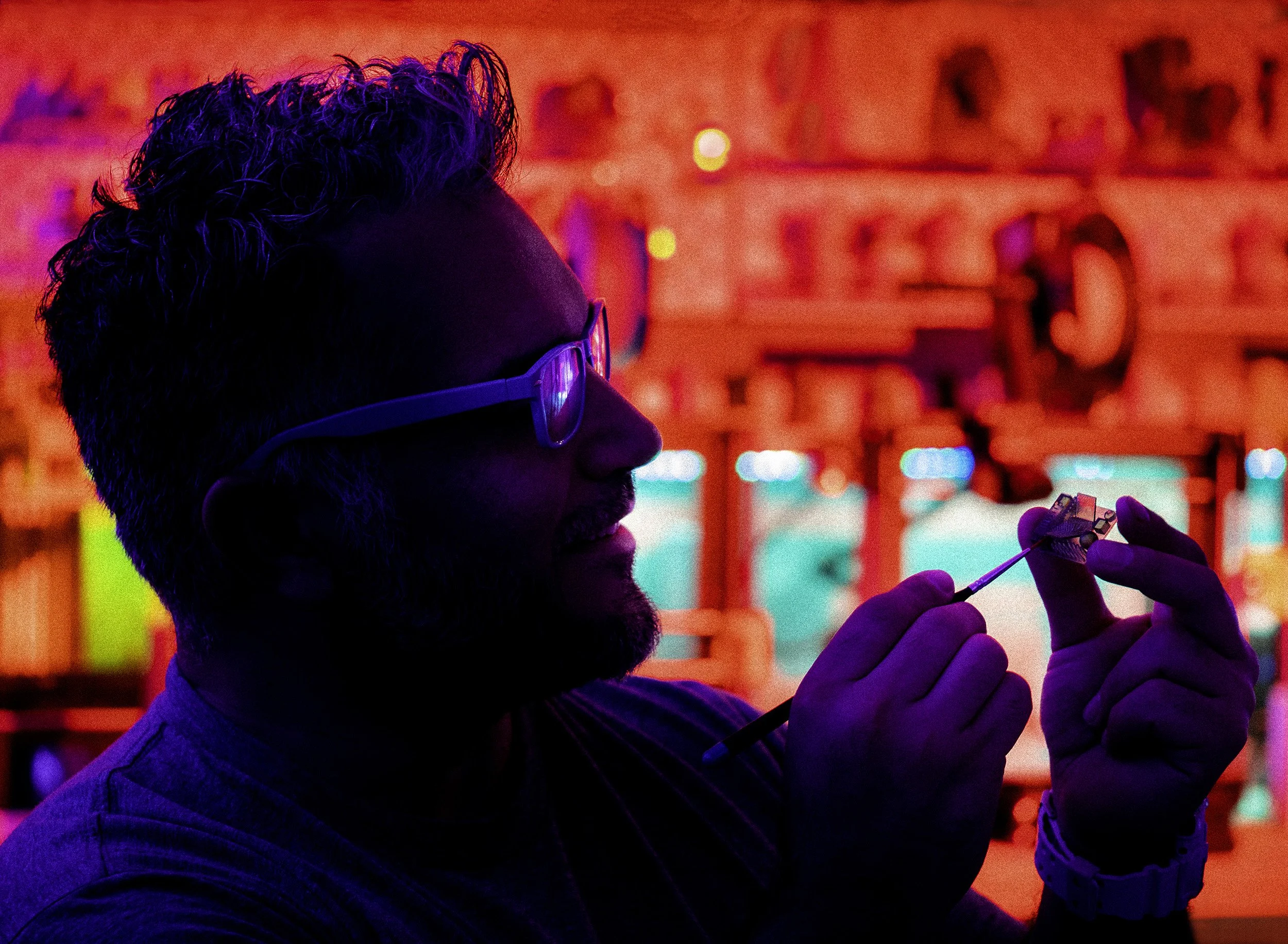 A man with curly hair and glasses holding a small object, possibly a microchip or electronic component, in a brightly colored environment with red, pink, and purple hues.