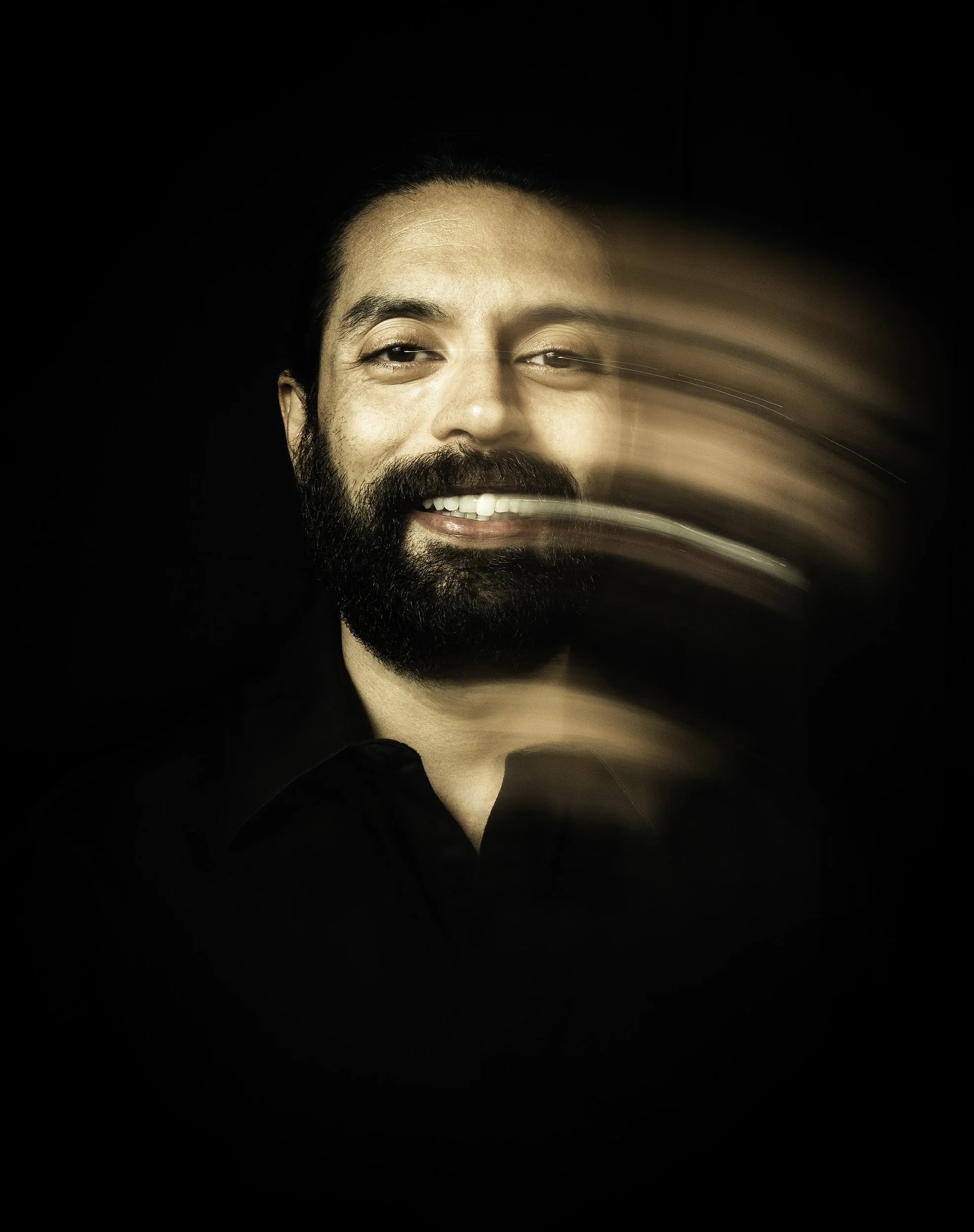 Close-up of a smiling man with a beard and mustache, his face partially blurred as if in motion, against a black background.