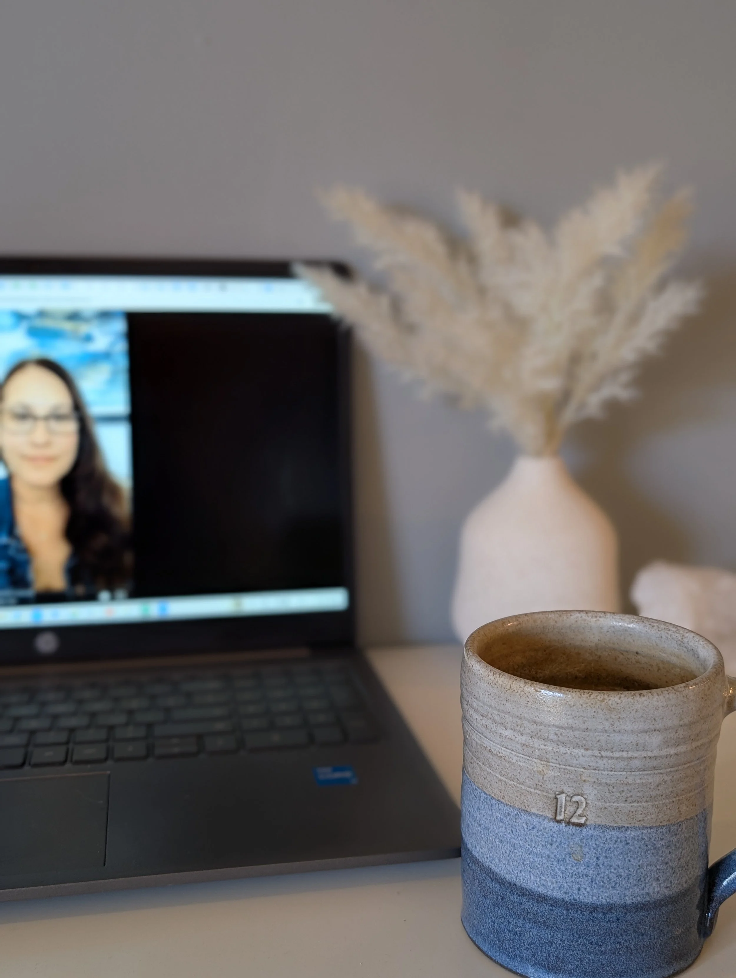 A ceramic mug filled with coffee, a laptop displaying a woman’s face on a video call, and a vase with dried pampas grass on a white surface.