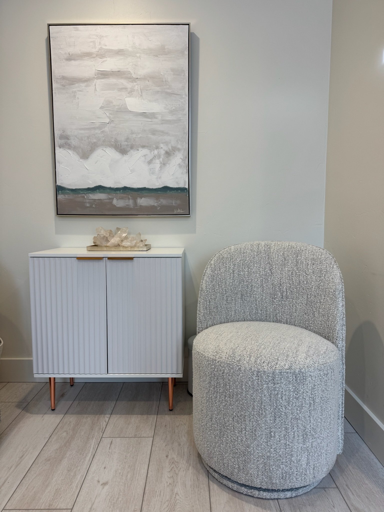 A modern interior corner featuring a white textured cabinet with gold handles and copper legs, topped with a crystal decor piece, a light gray upholstered circular chair with a curved back, a large abstract painting in shades of gray and green, and light gray wood plank flooring.