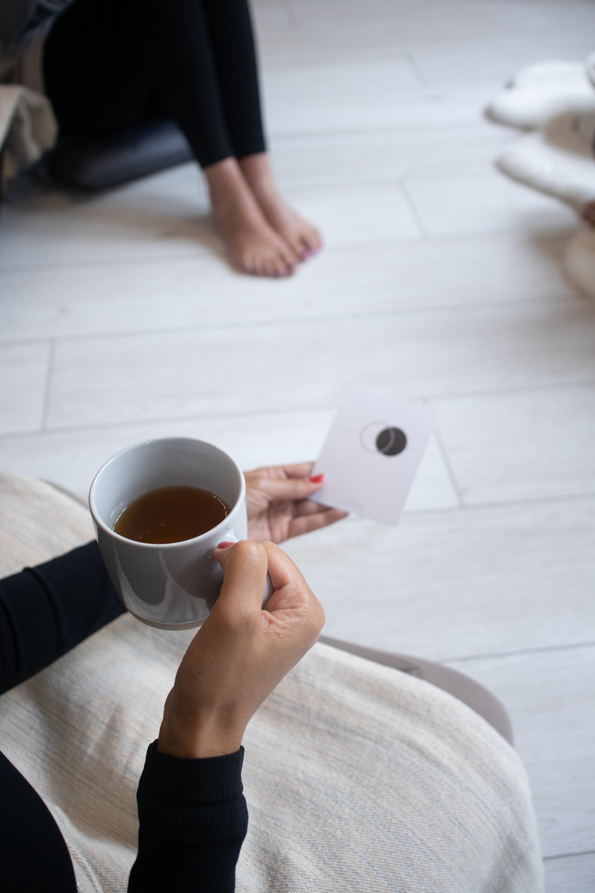 Close-up of a person's hand holding a white mug with hot tea, and another hand holding a small paper card with a black circle design, with a woman seated nearby and her feet visible on a white wooden floor.