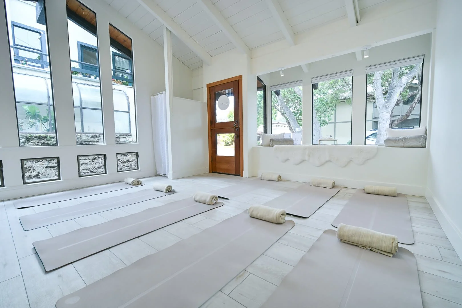 Yoga class setup with mats and rolled towels in a bright, airy room with large windows and trees outside.