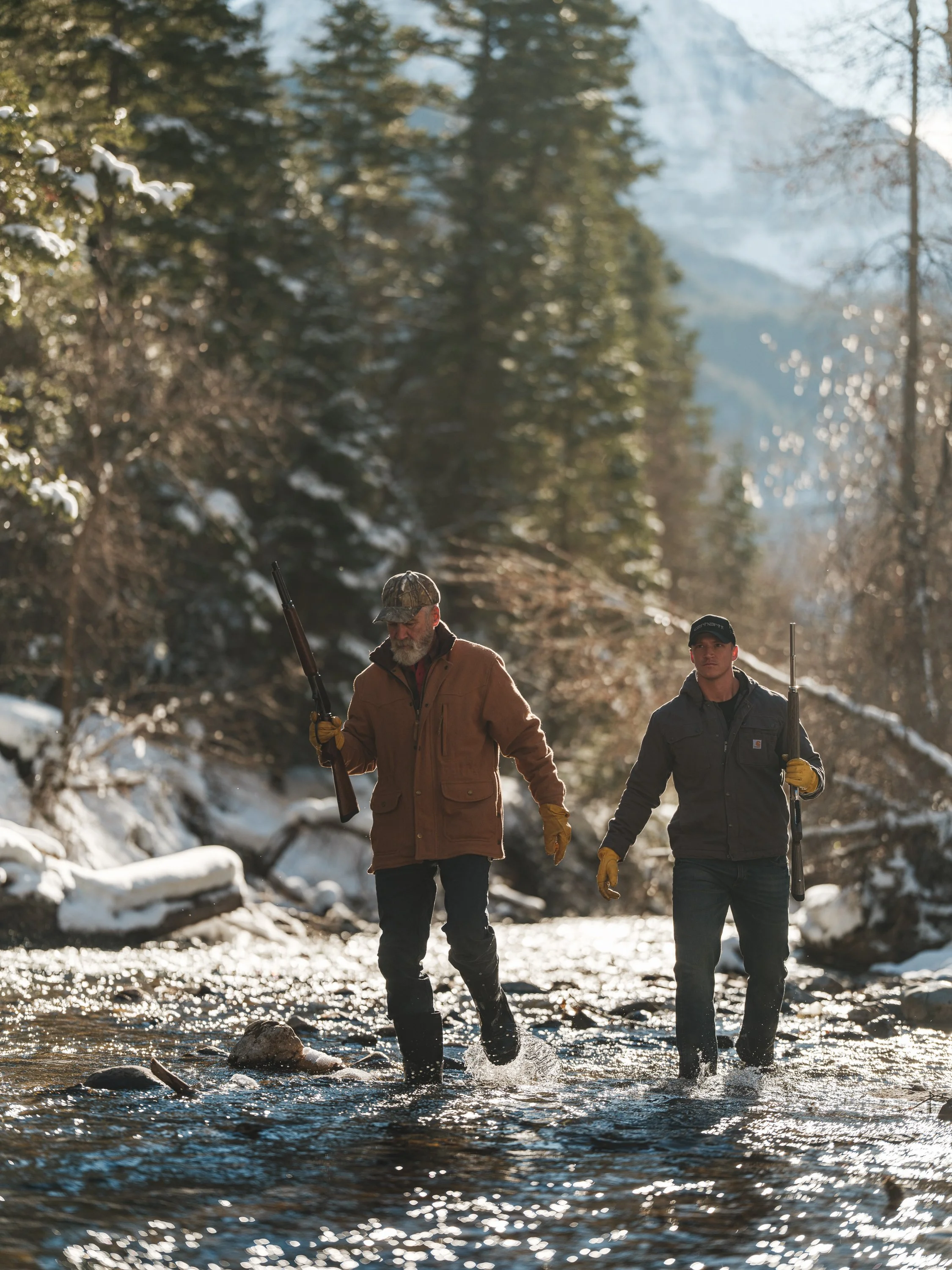 Two people wearing outdoor clothing wading through a shallow, snowy creek, carrying shotguns, with a forested mountain backdrop.