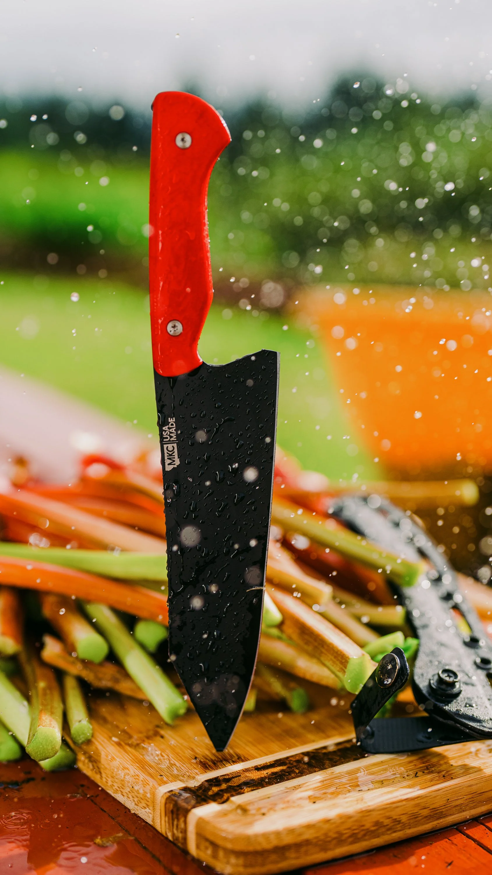 Close-up of a black-bladed knife with a red handle standing upright on a cutting board with rhubarb and sprinkled water droplets.
