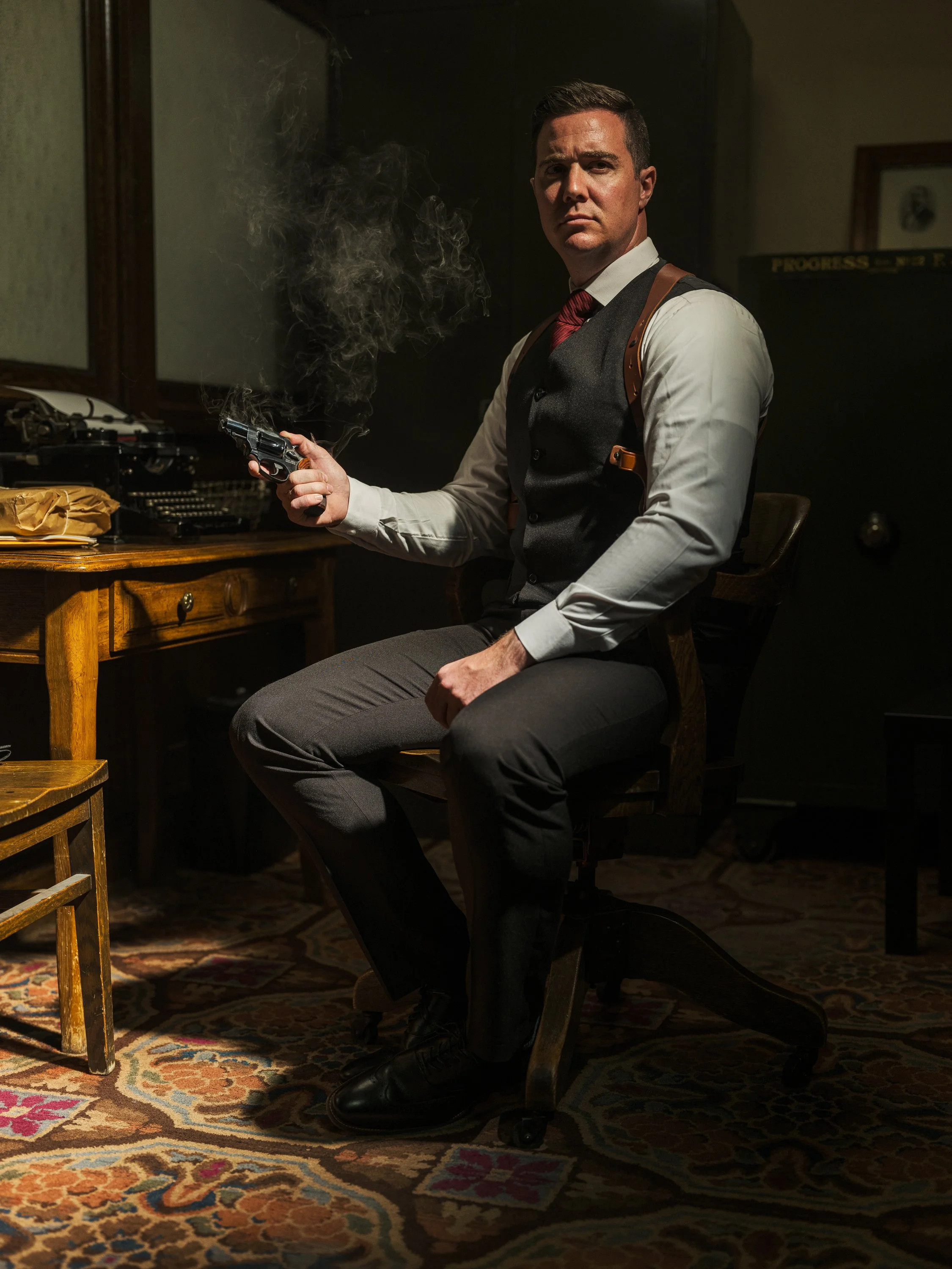 Man in vintage office setting holding a smoking gun, wearing a vest and shoulder holster, seated at a desk with a typewriter, in low light ambiance.