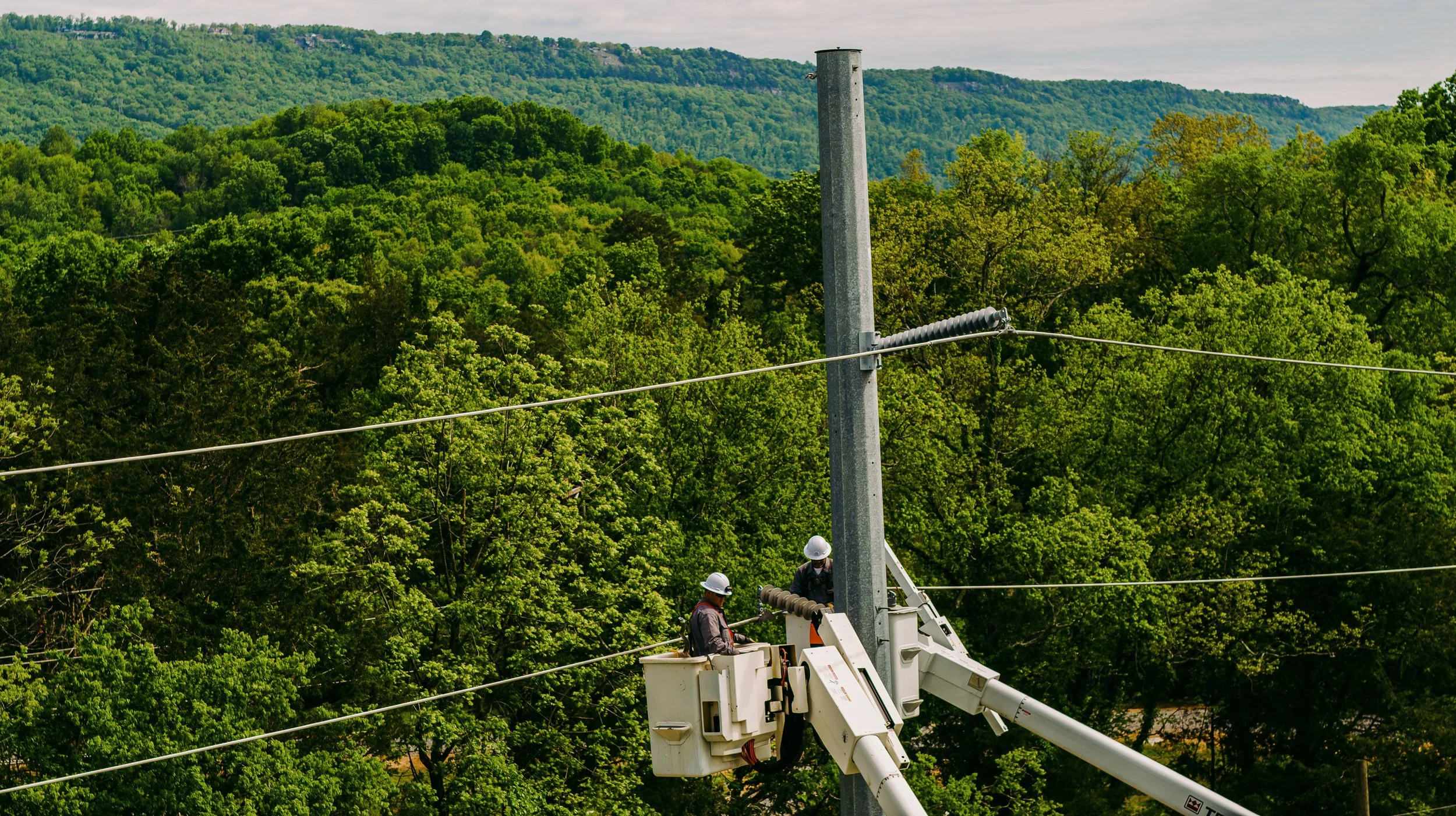 Two workers in a bucket truck repairing power lines in a forested area.
