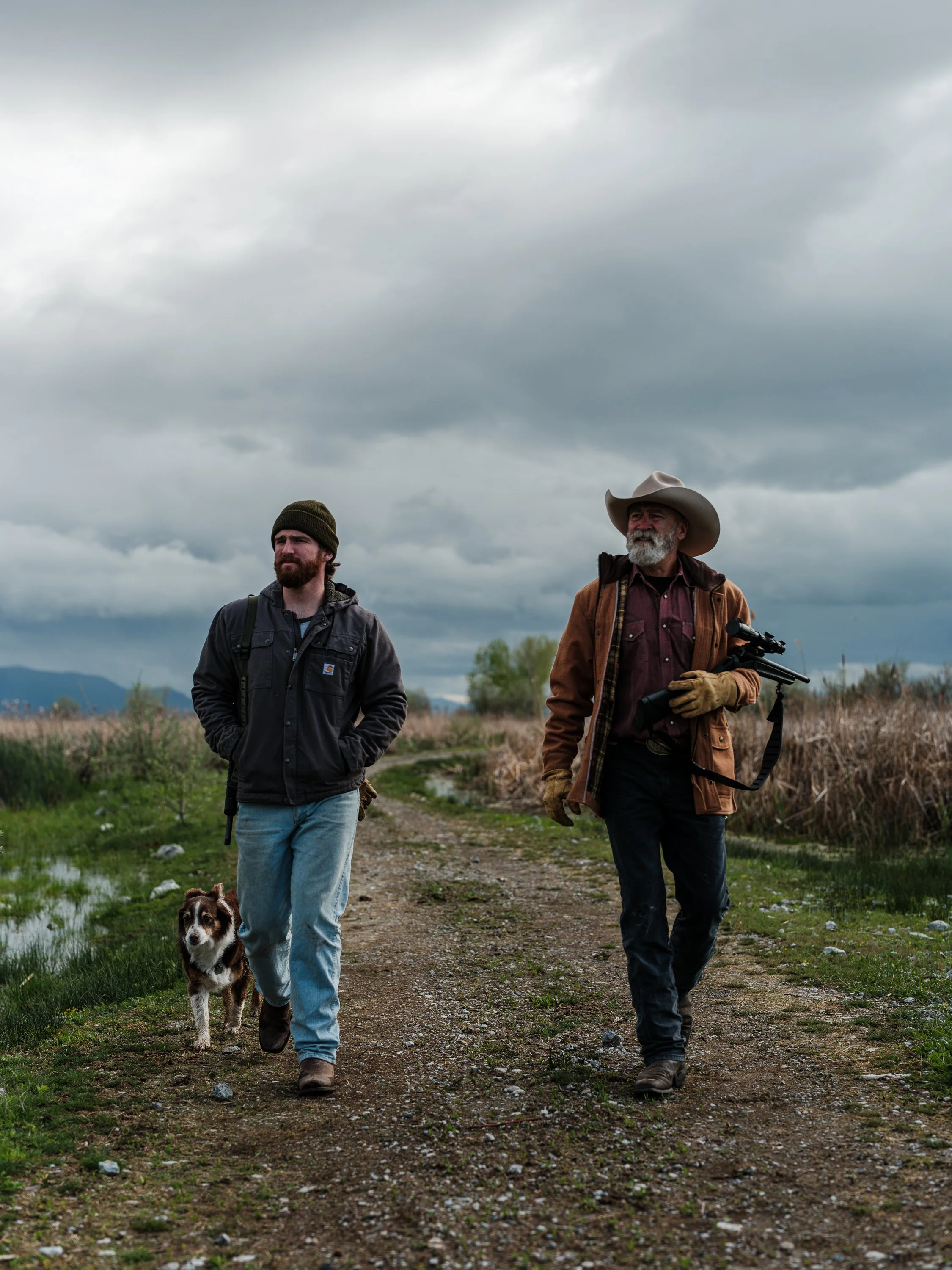 Two men wearing outerwear and hats walking on a rural path with a dog, under a cloudy sky.