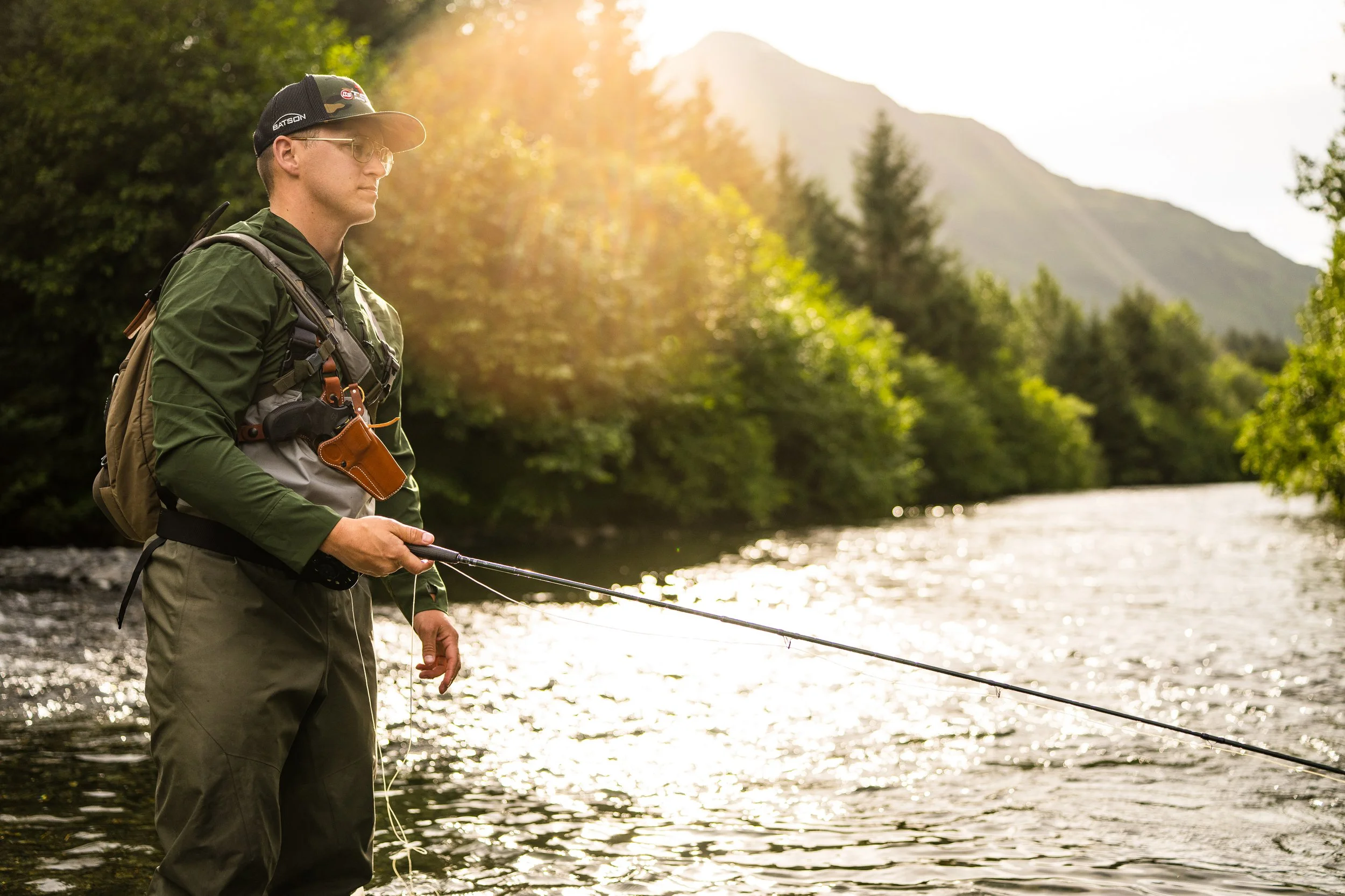 Man fly fishing in a river with sunlit forest and mountains in the background.