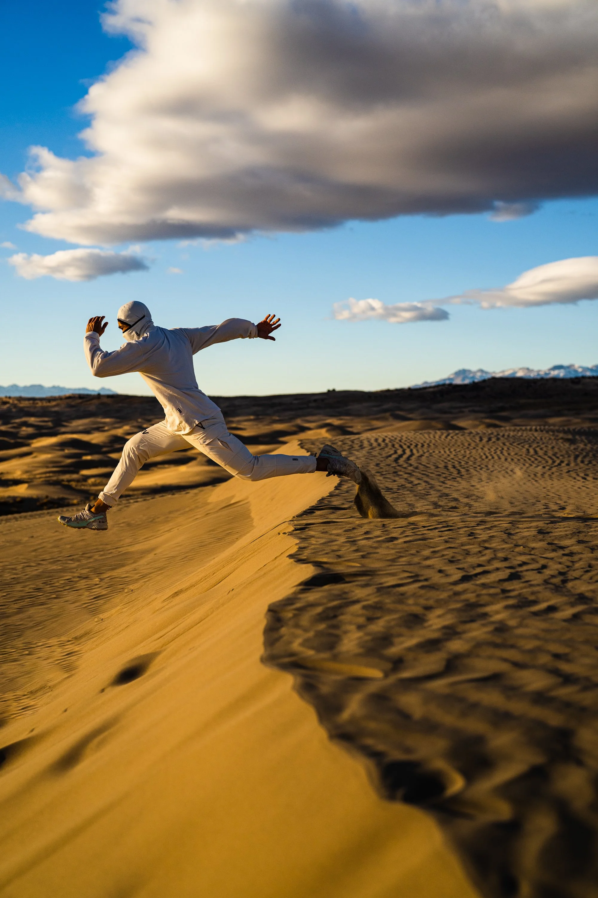 Person in white clothes jumping over sand dune in desert with cloudy sky.
