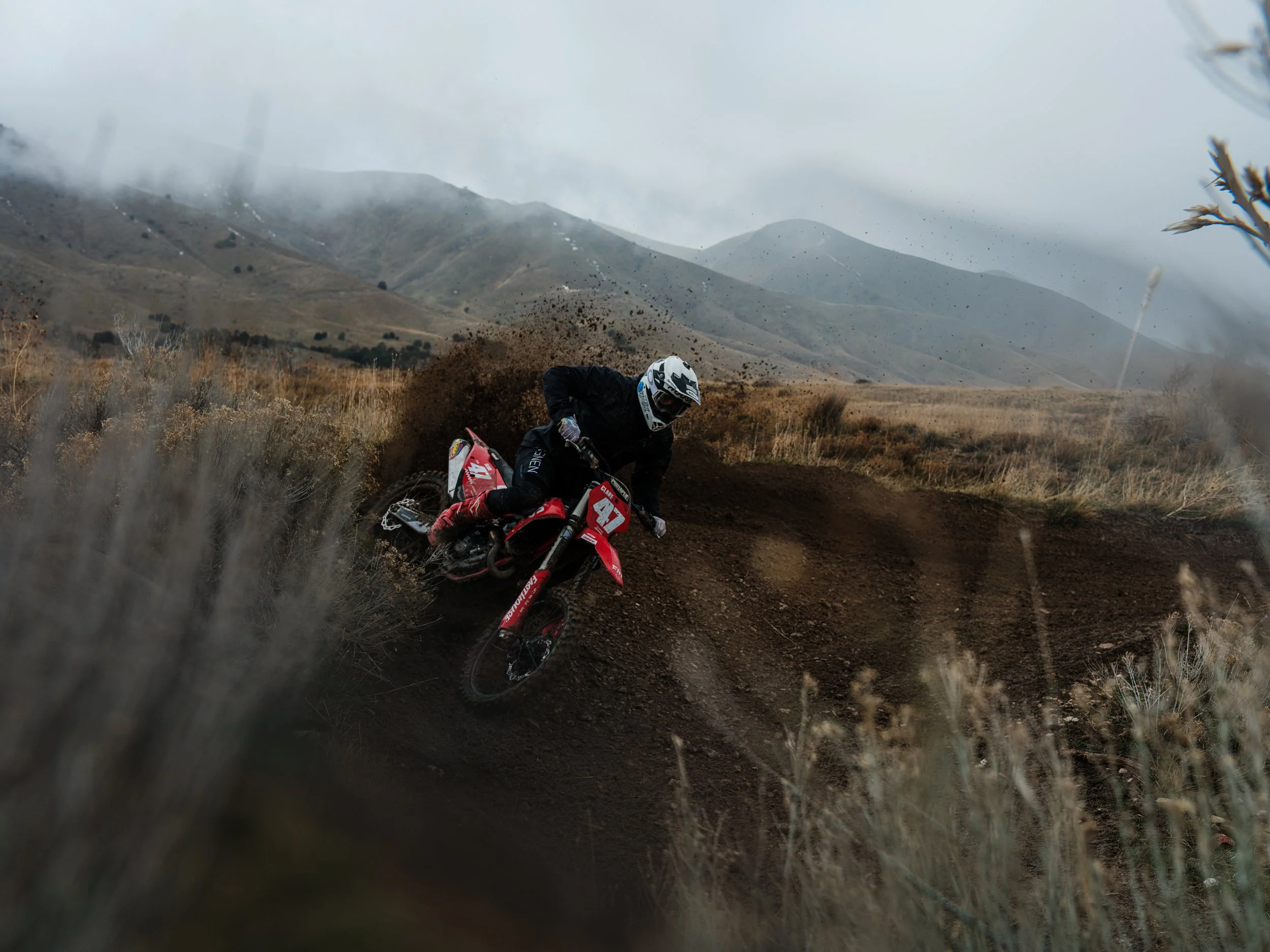 Dirt bike rider making a sharp turn, kicking up dirt, in a rugged landscape with mountains and cloudy skies in the background.