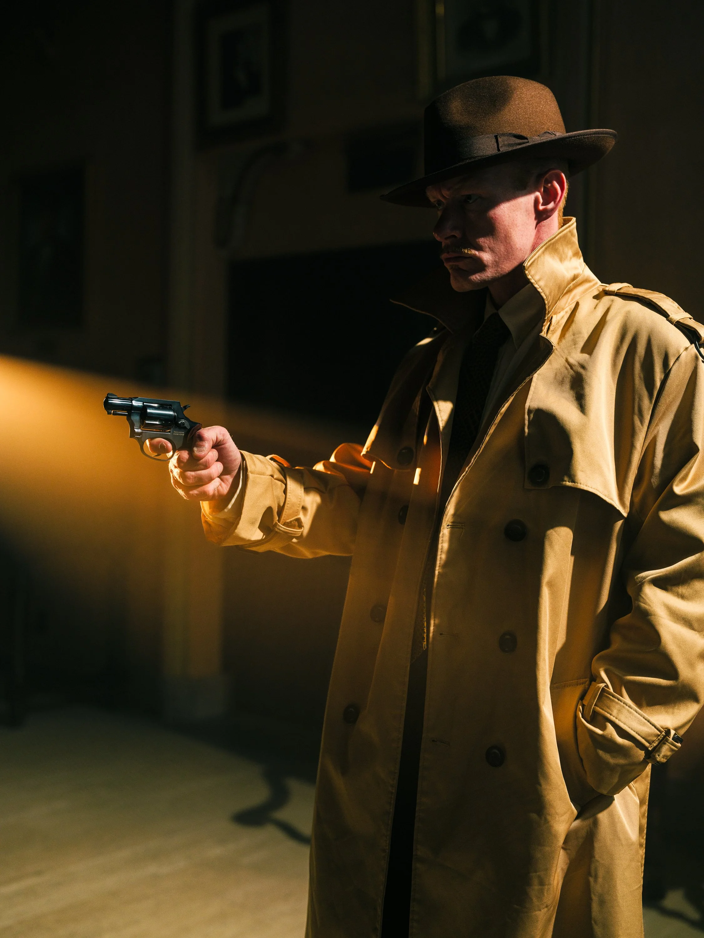 Detective in trench coat and fedora holding a revolver, standing in dramatic lighting.