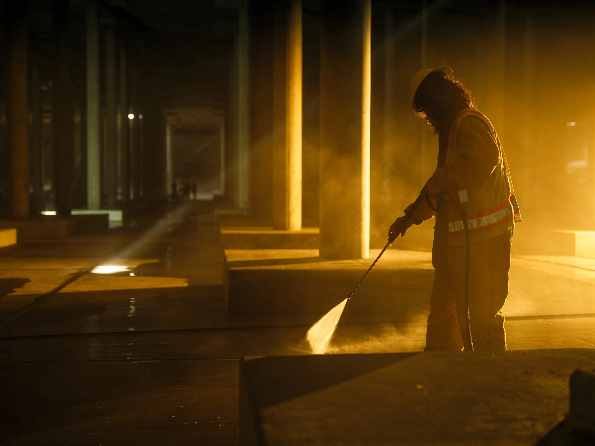 Person using a pressure washer in a dimly lit industrial space with concrete pillars and beams.