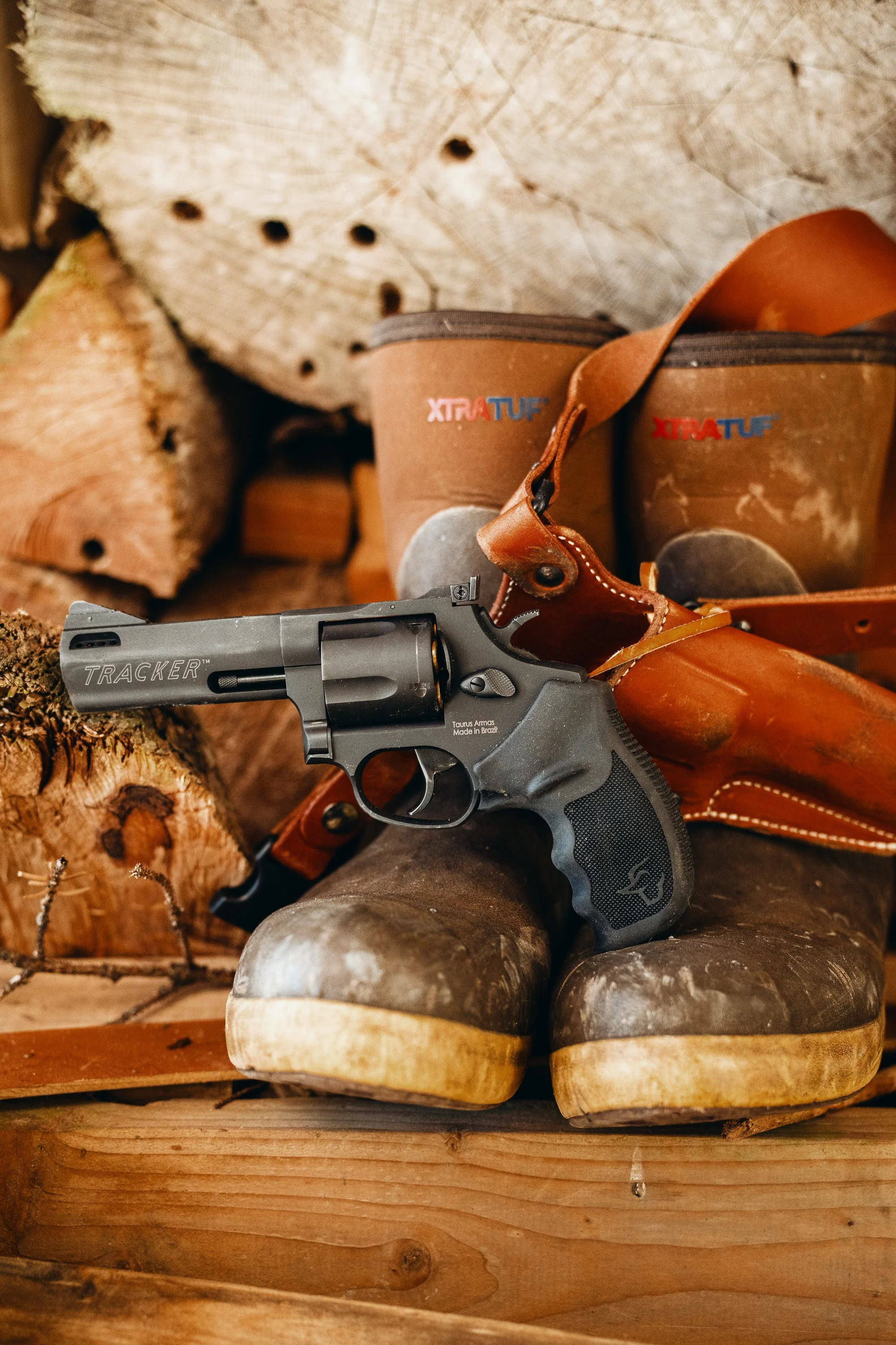 Close-up of a revolver with a leather holster resting on muddy boots in front of stacked wood logs.