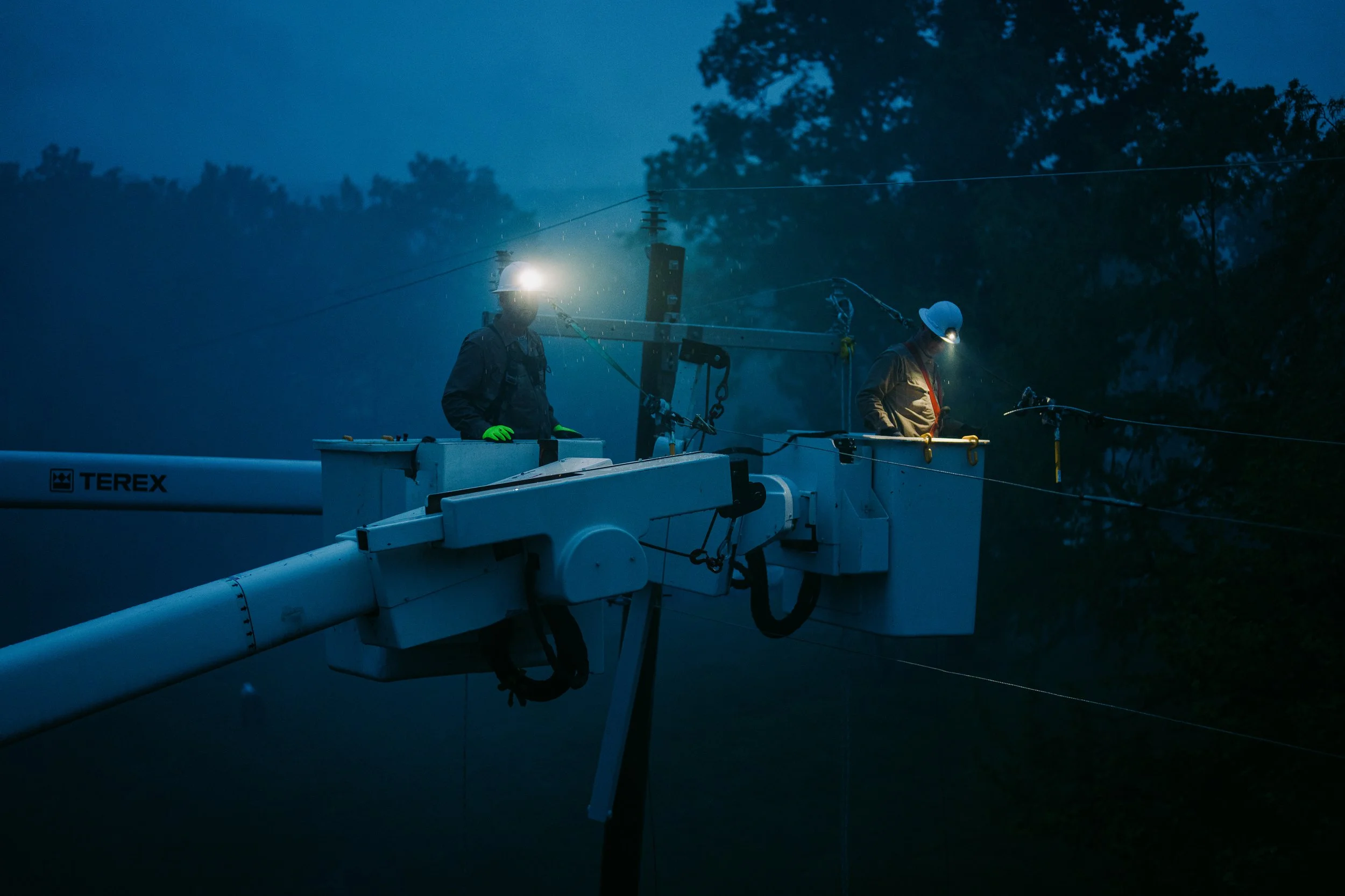 Utility workers in bucket trucks repairing power lines at night with headlamps.