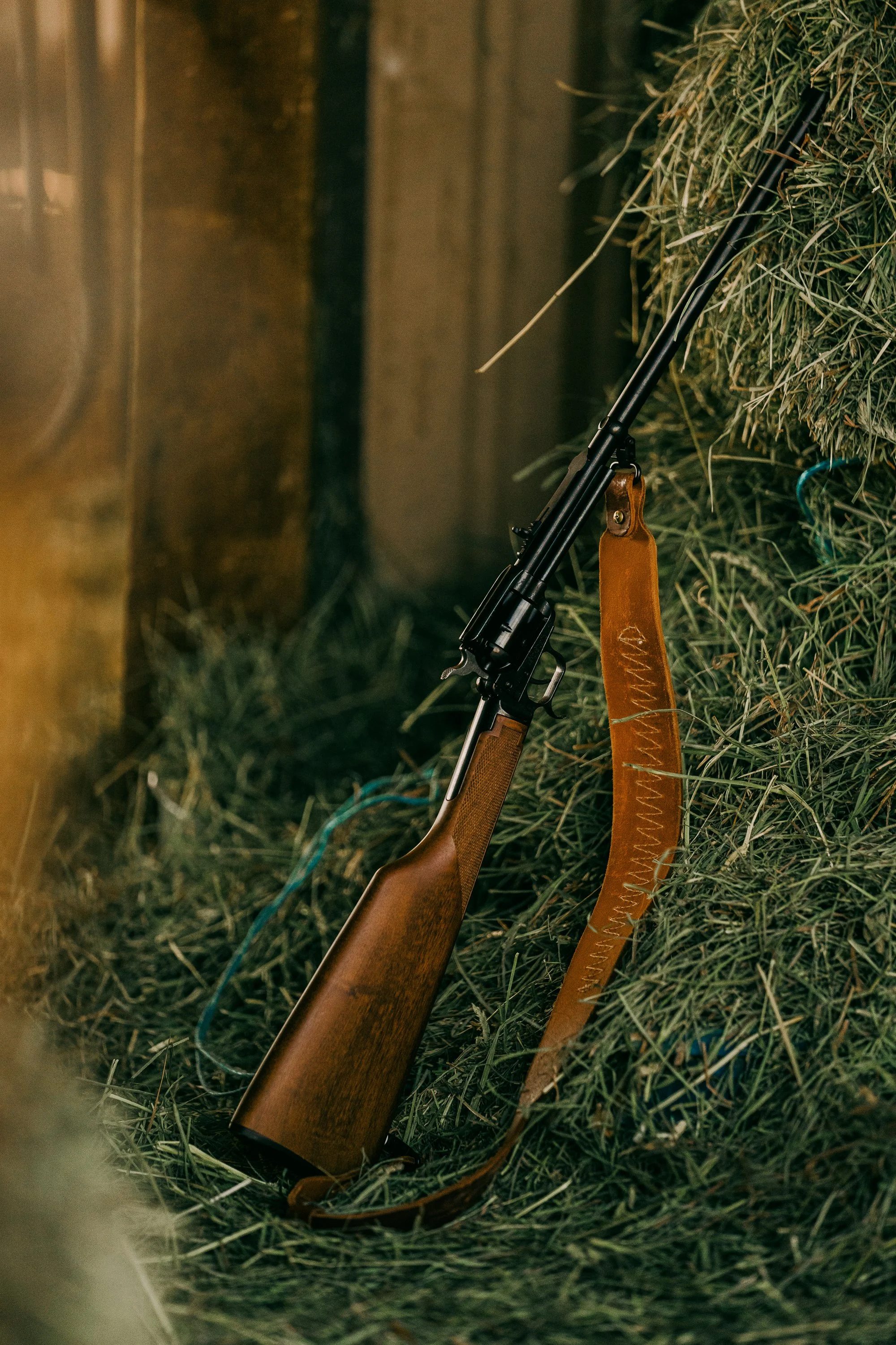 A rifle with a wooden stock and leather sling leaning against hay bales in a rustic setting.