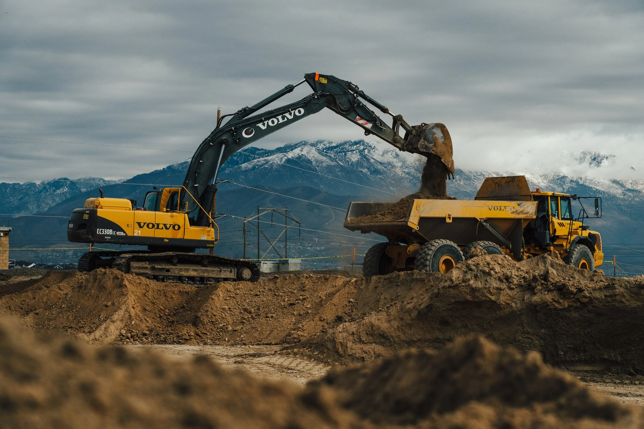 A construction site with a Volvo excavator loading dirt into a Volvo dump truck against a mountainous backdrop.