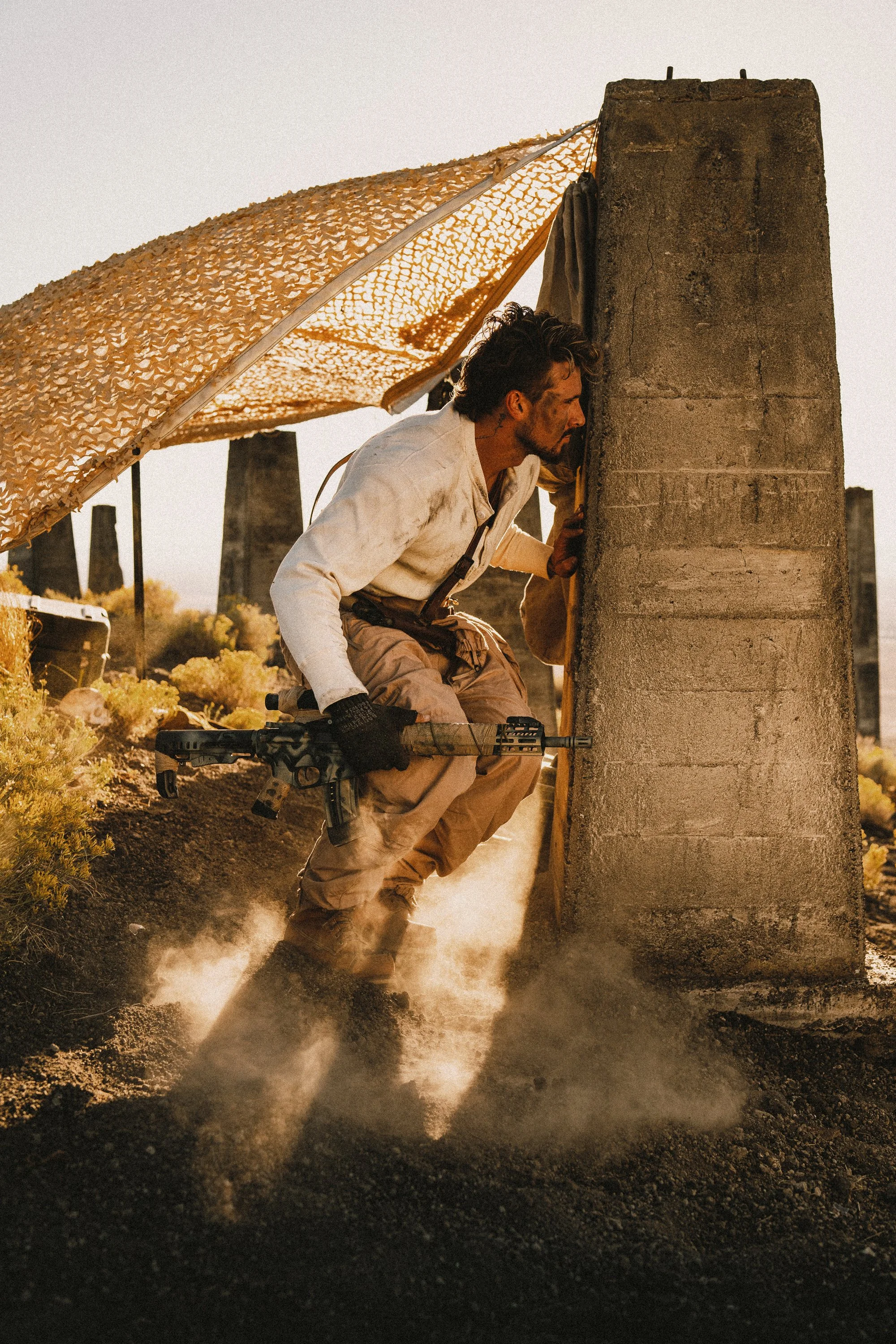 Man in tactical gear crouching and peeking around a concrete pillar with a rifle in a desert setting.