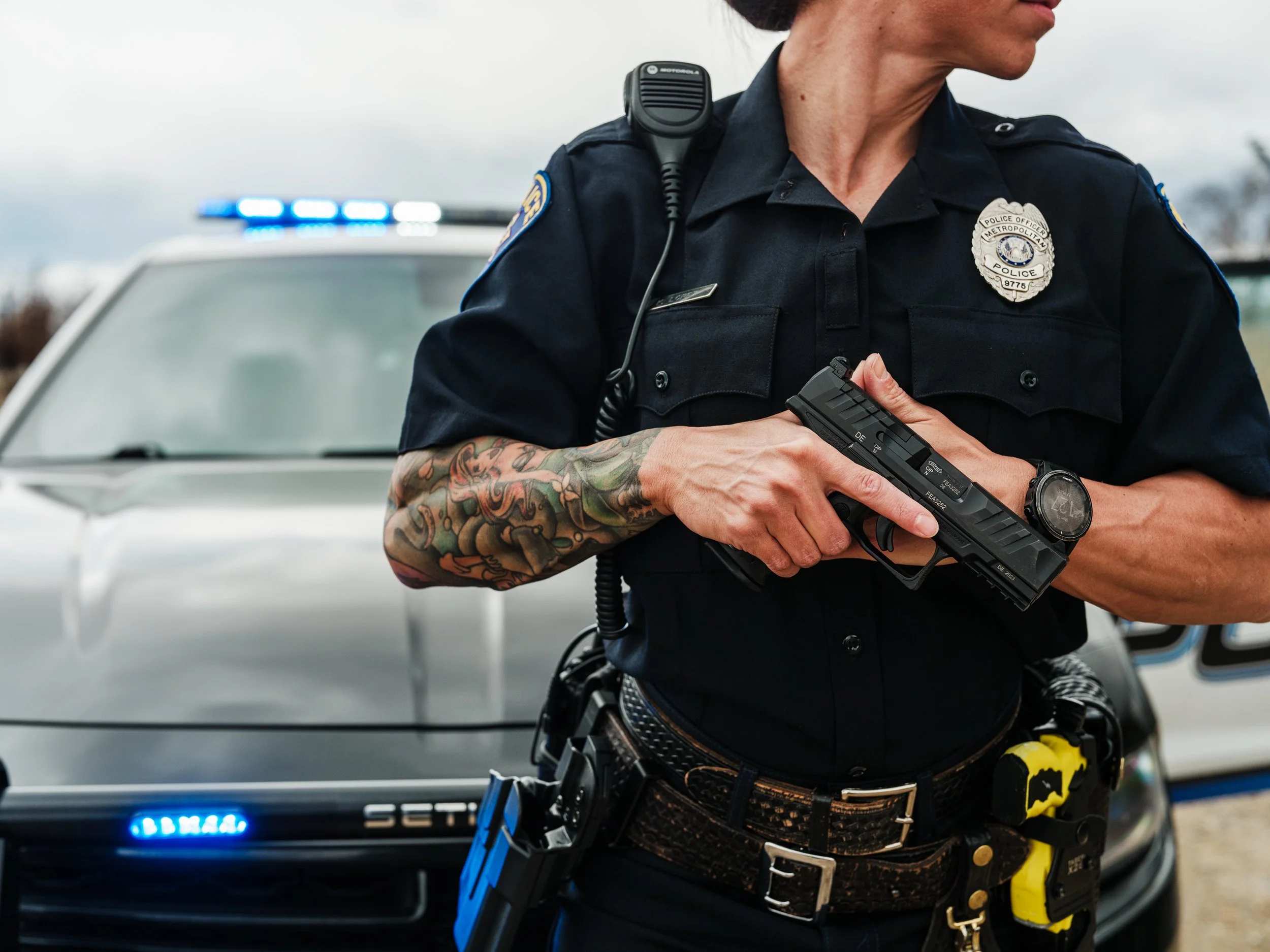 Police officer standing in front of a patrol car with a handgun in hand and a tattoo sleeve on their arm.