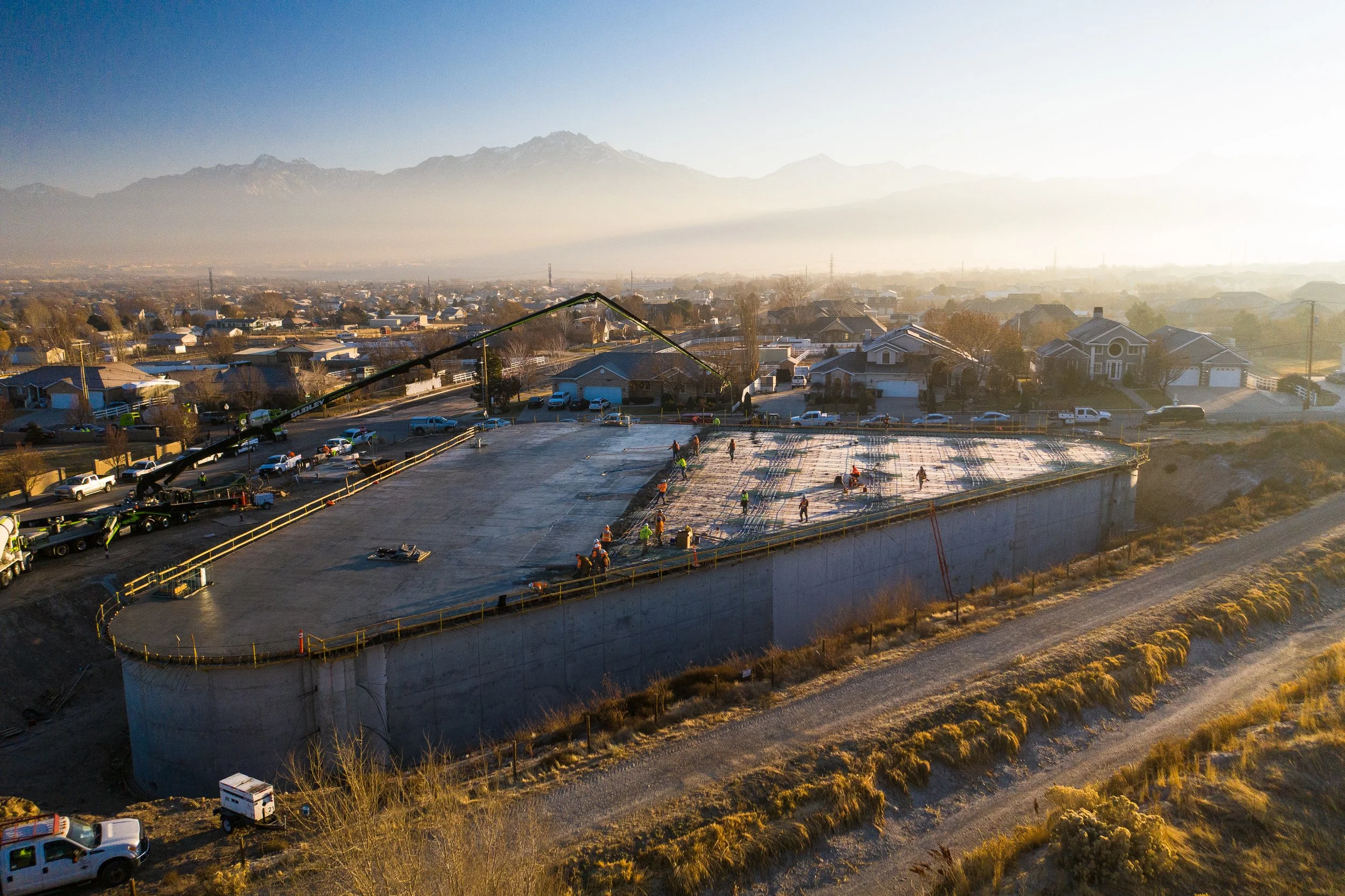 Aerial view of a large concrete construction site with workers and machinery, surrounded by residential houses and mountains in the background.