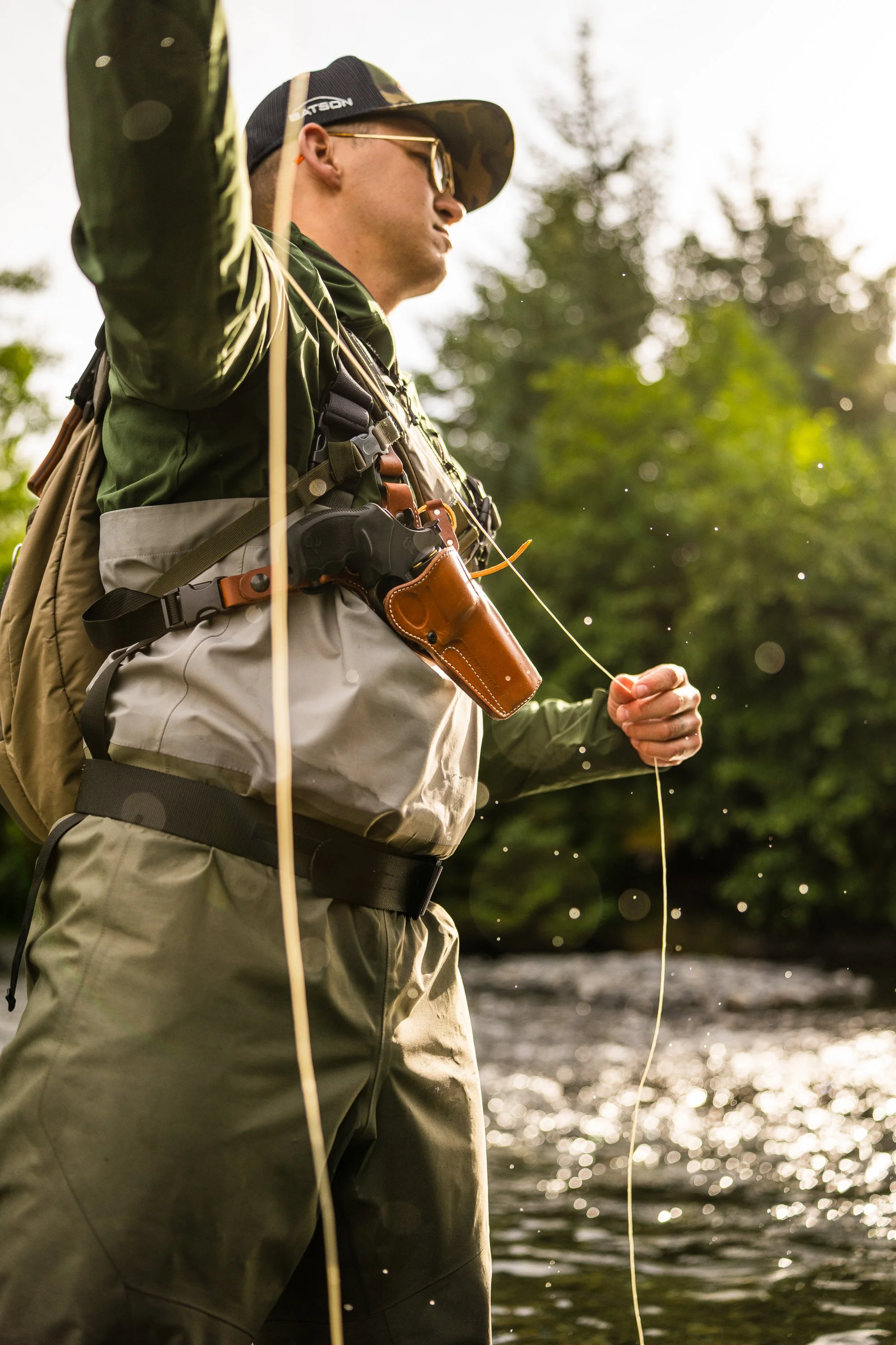 Person fly fishing in a river, wearing waders, a cap, and a holstered tool. Background of trees and water.