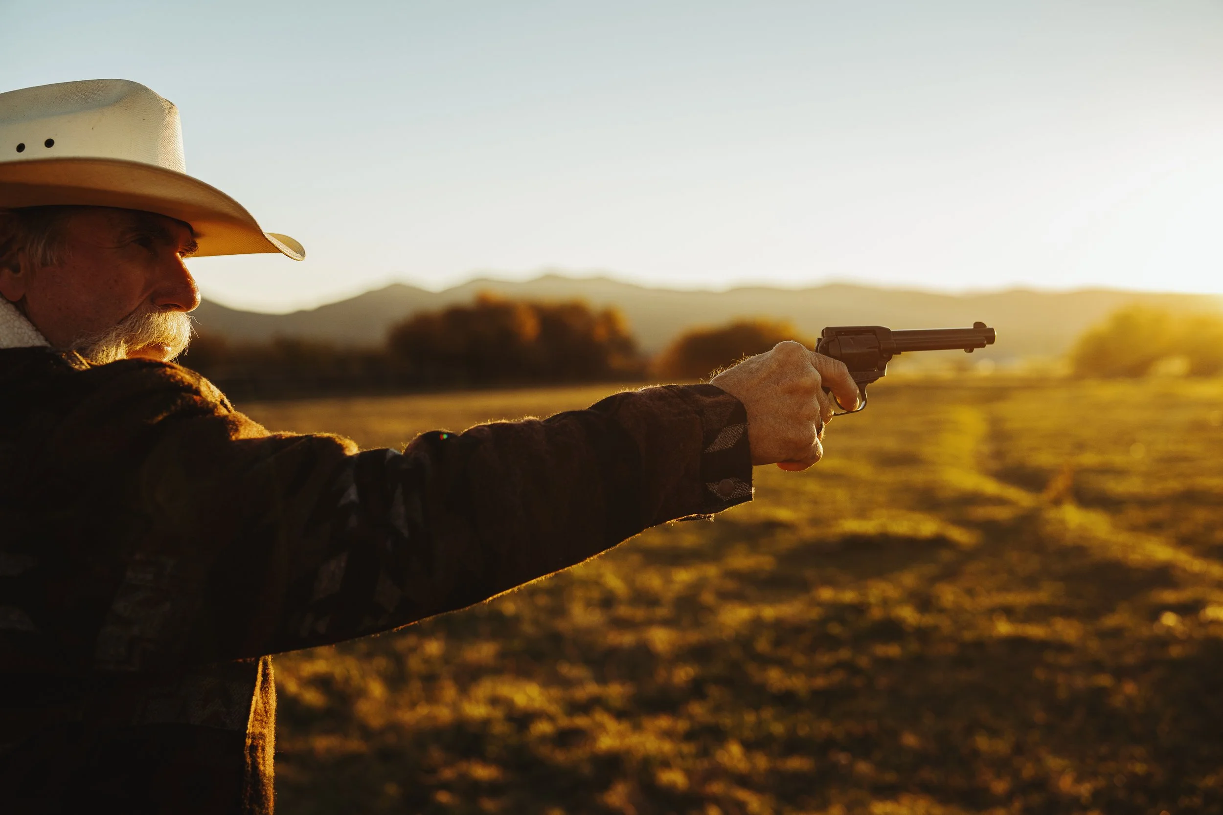 Man wearing cowboy hat aiming a revolver outdoors at sunset.